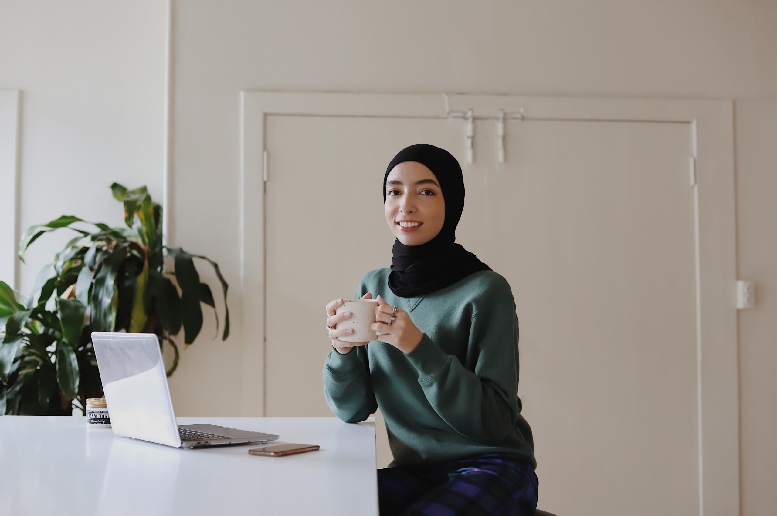 en ung kvinne sitter ved et bord foran en pc, hun holder en kopp