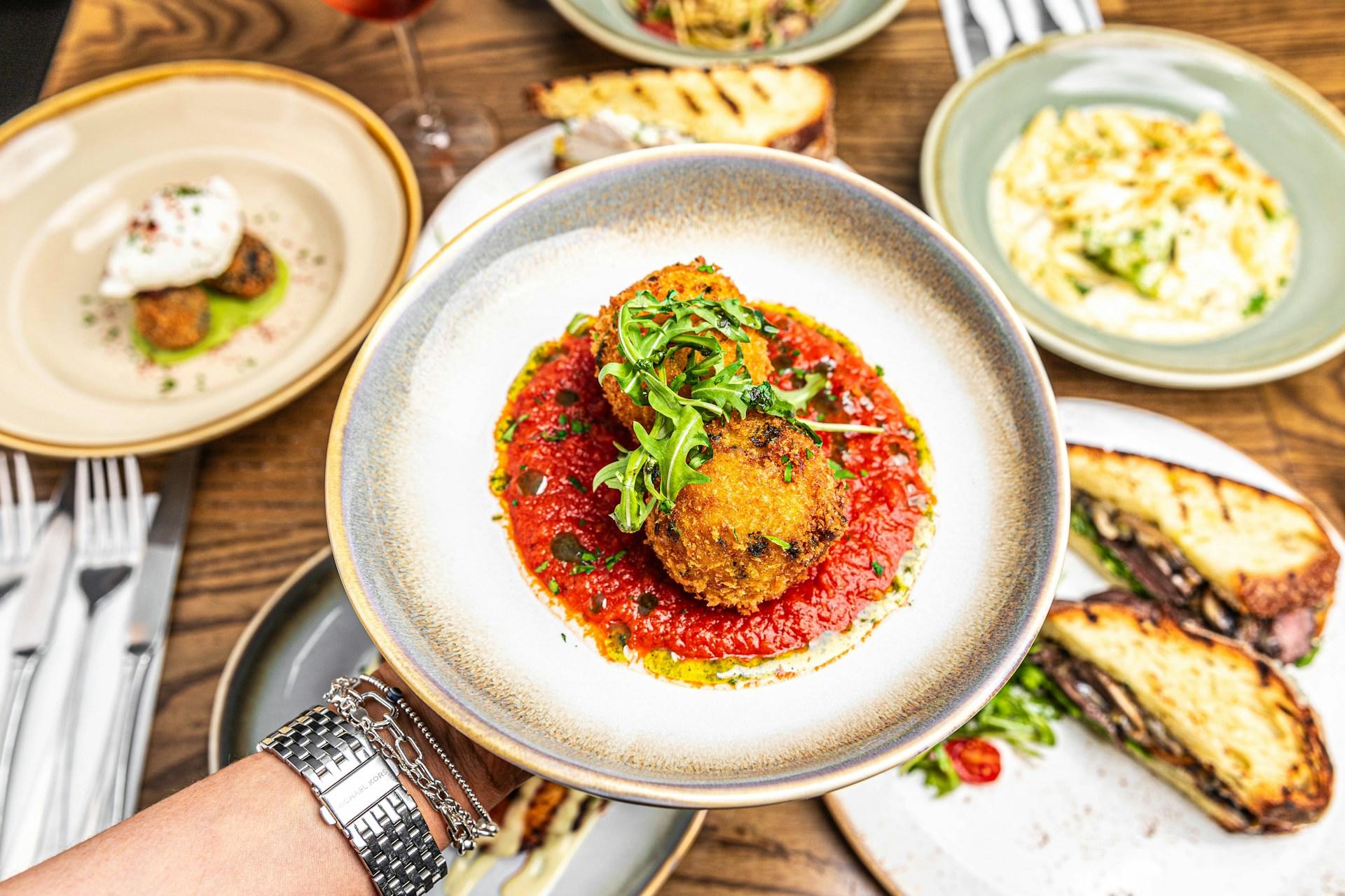 Hånd holder tallerken med arancini på, men andre retter i bakgrunnen.