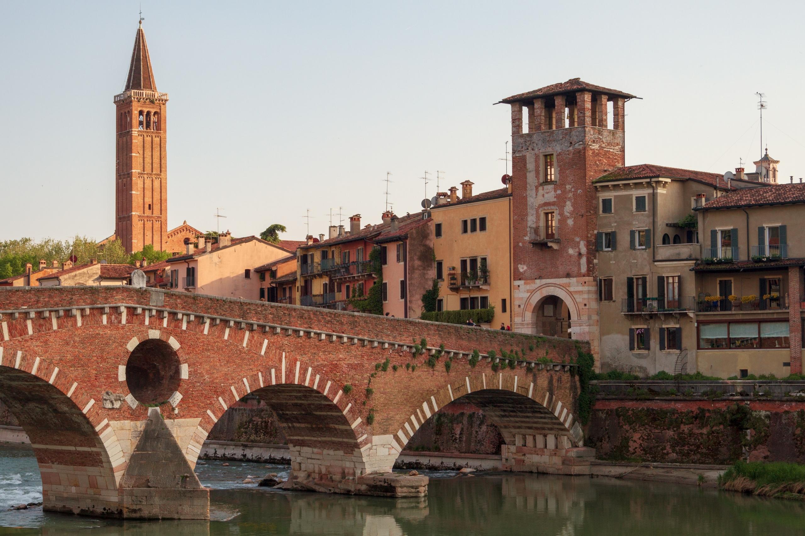 puente pietra i verona, en bro over en elv med brune, slitte hus i bakgrunnen og tårn i bakgrunnen