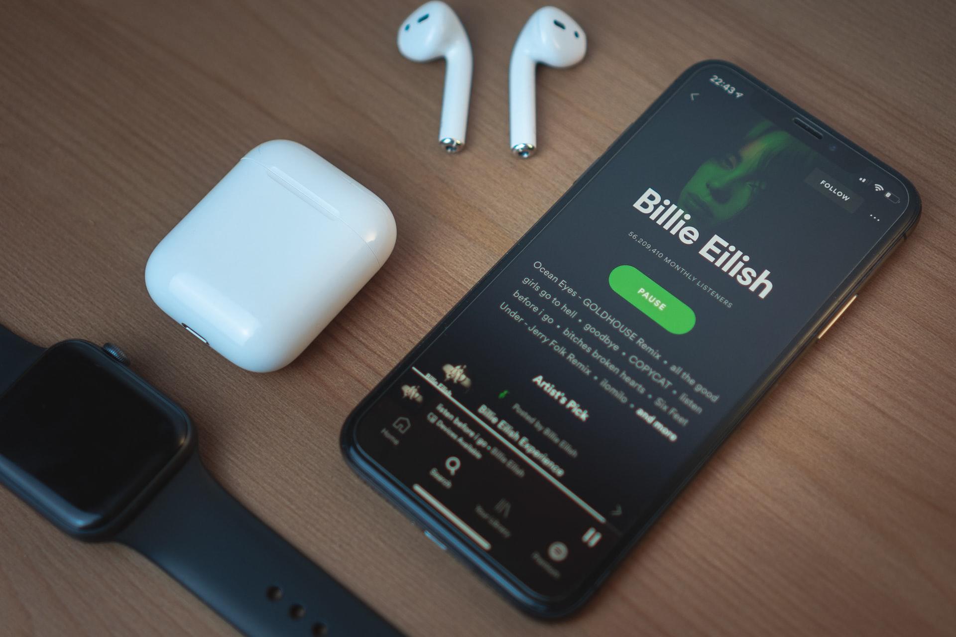 Sort iPhone med Billie Eilish på Spotify, og Apple Watch, Airpods og etui ligger ved siden av.