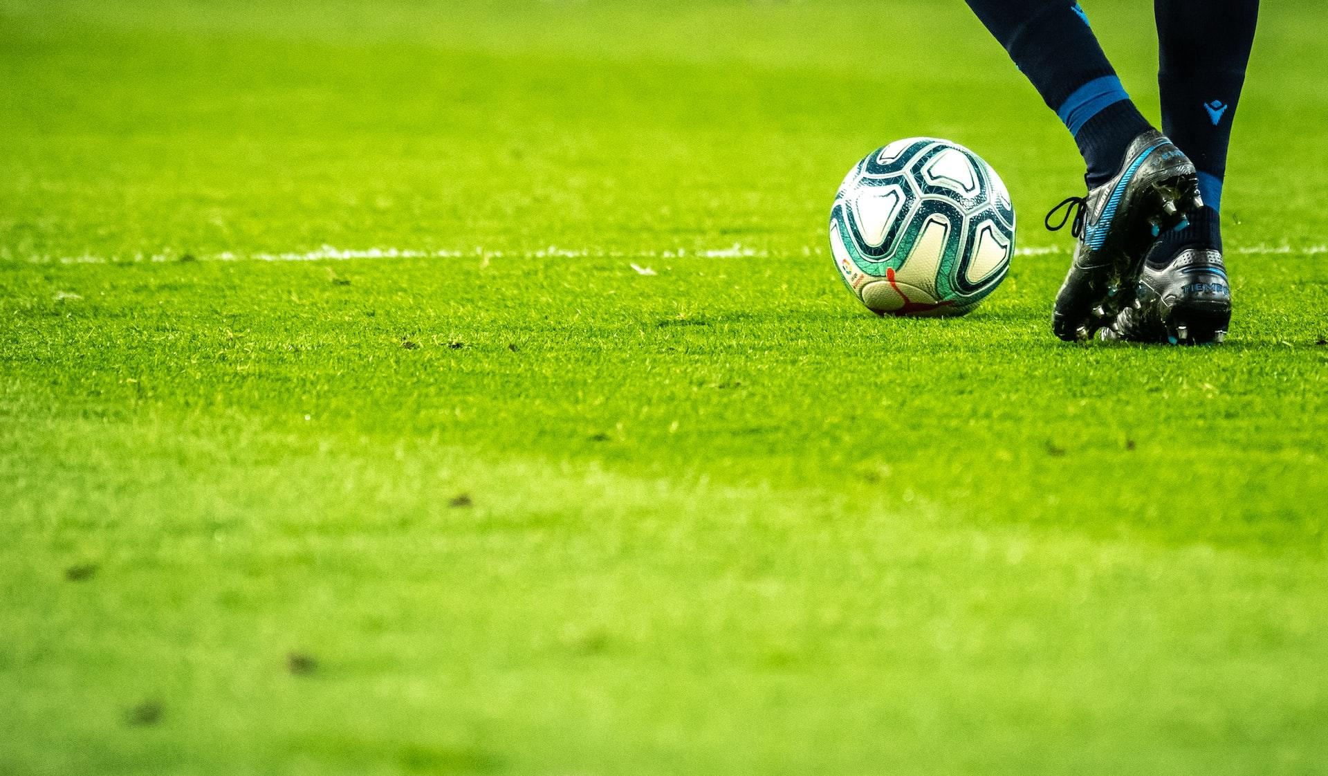 Fotball med mønster triller bortover gress mens føtter med mørke fotballsko sparker etter ballen.