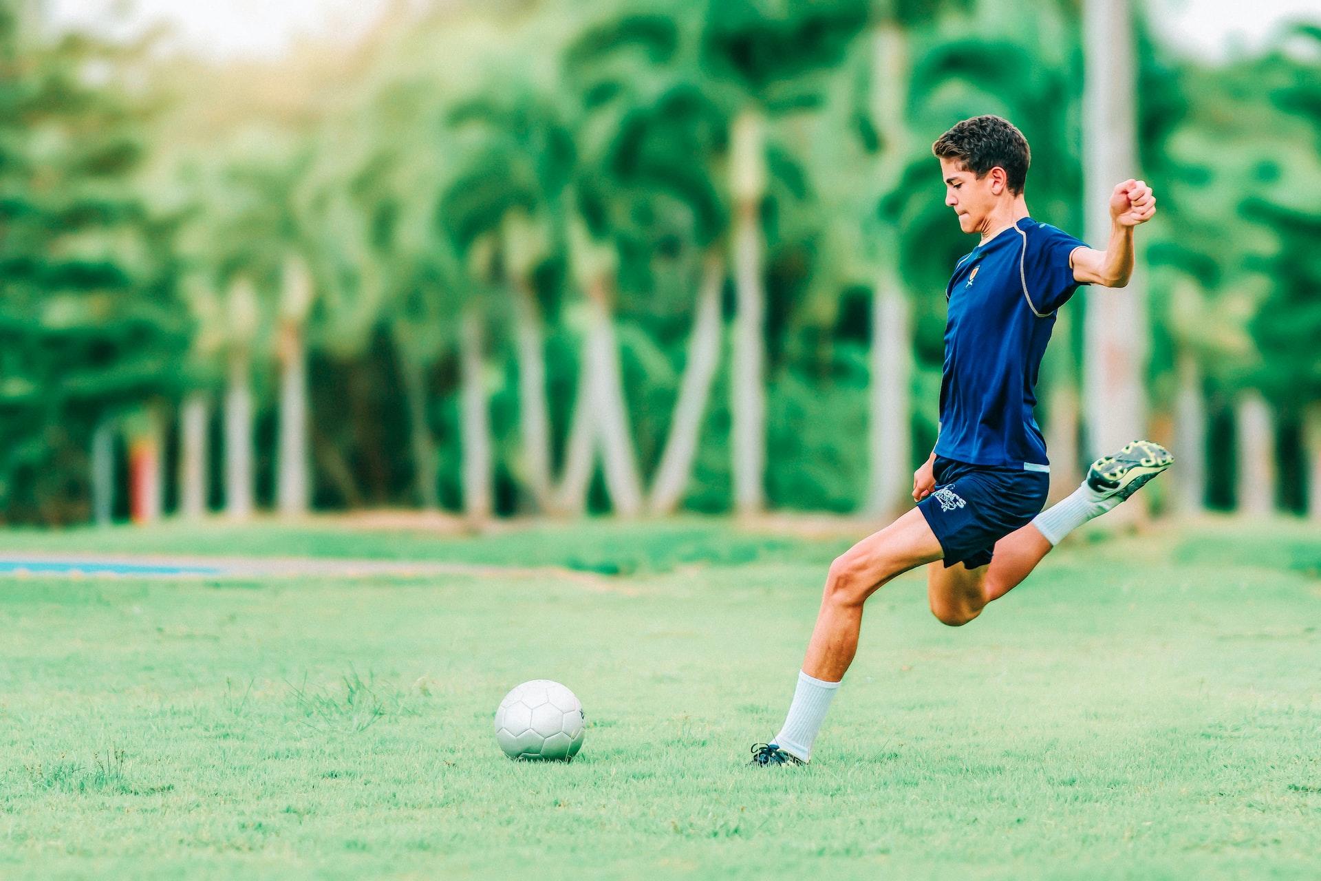 Gutt med brunt hår og mørkeblå fotballdrakt skal til å sparke en fotball i fart ute på en gressplen med trær i bakgrunnen.