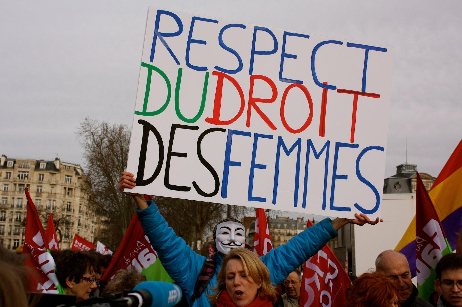 En person med maske holder et stort skilt med teksten "Respect du droit des femmes" i en mengde med mennesker.