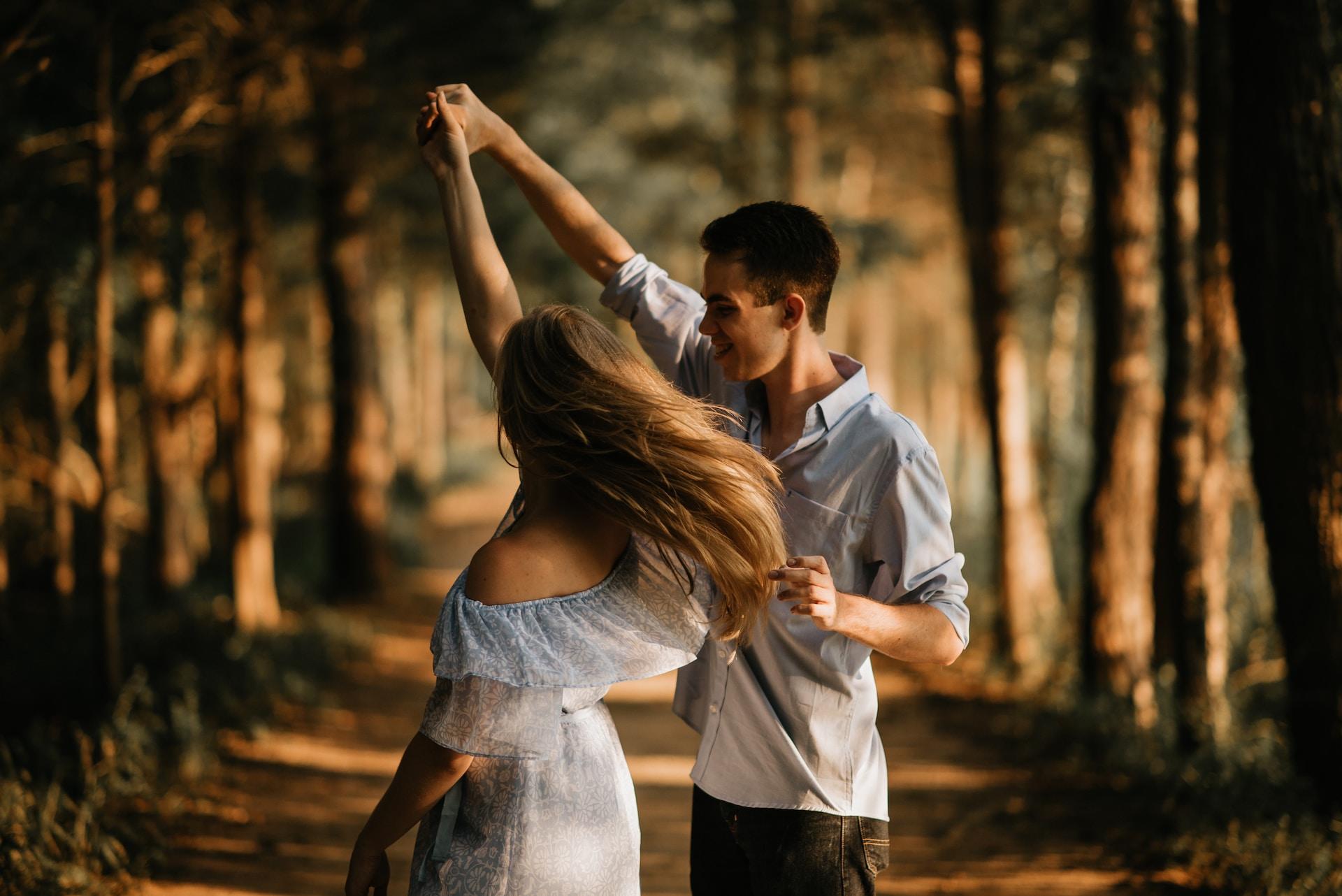 Ung kvinne med lyseblå kjole og langt lyst hår danser sammen med ung mann med olabukser og lyseblå skjorte i skogen.