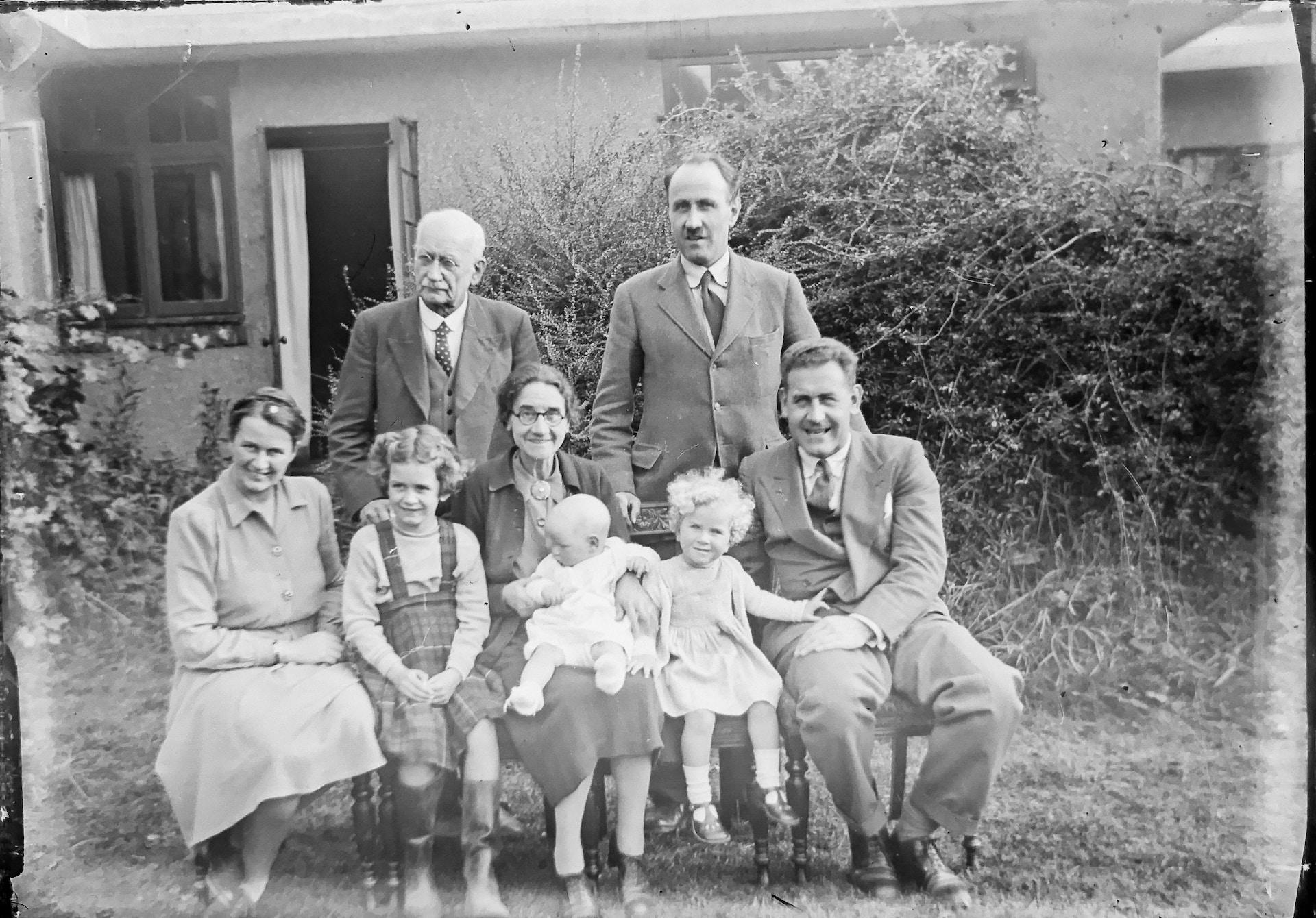 Gammelt bilde i svart-hvitt viser en familie som blir fotografert utenfor huset sitt, bestående av åtte familiemedlemmer i alle aldre.