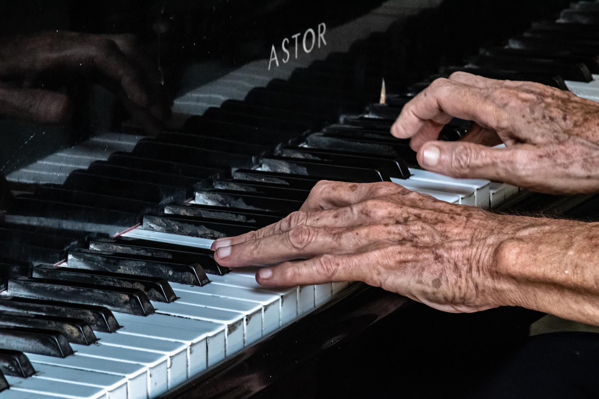 Eldre hender med føflekker og rynker spiller på et slitt piano.