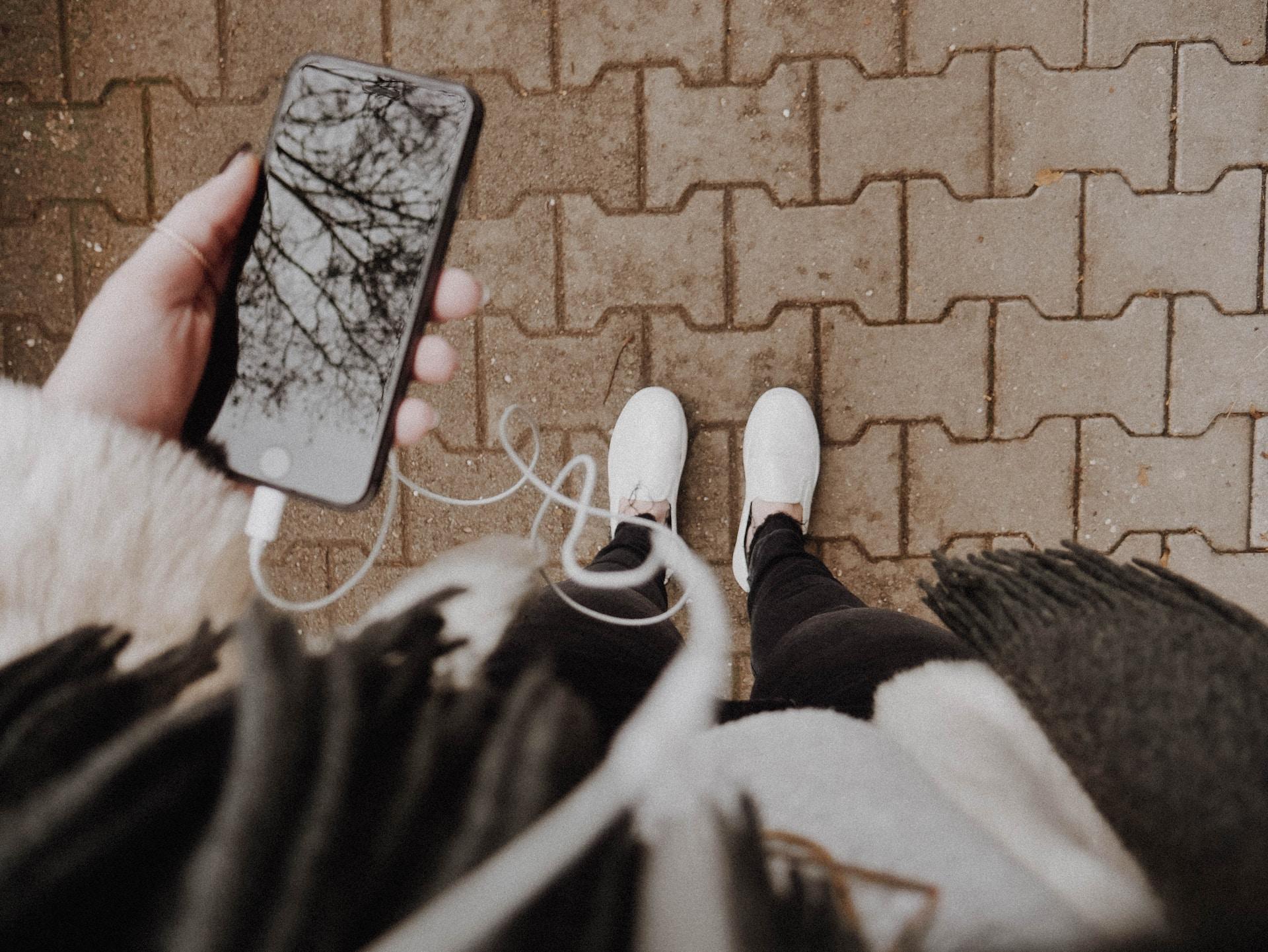 Bilde tatt ovenfra av en jente med hvite sko, sorte jeans, hvit genser or grått skjerf som hører på musikk fra iphonen sin med ørepropper plugget i.