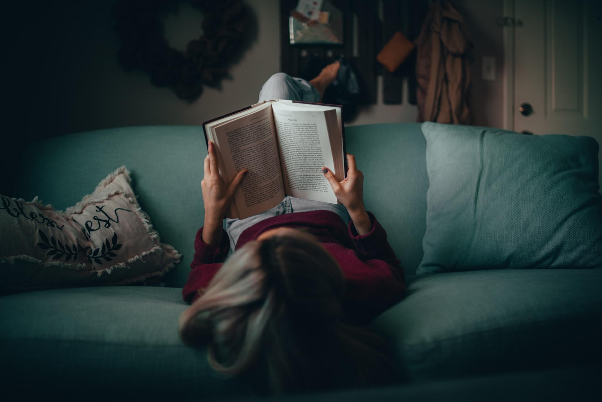 Dame med rød genser og jeans ligger opp ned på en grønn sofa og leser i en bok.