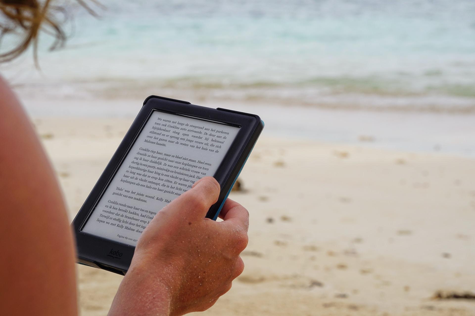 Person leser bok på nettbrett på en strand.