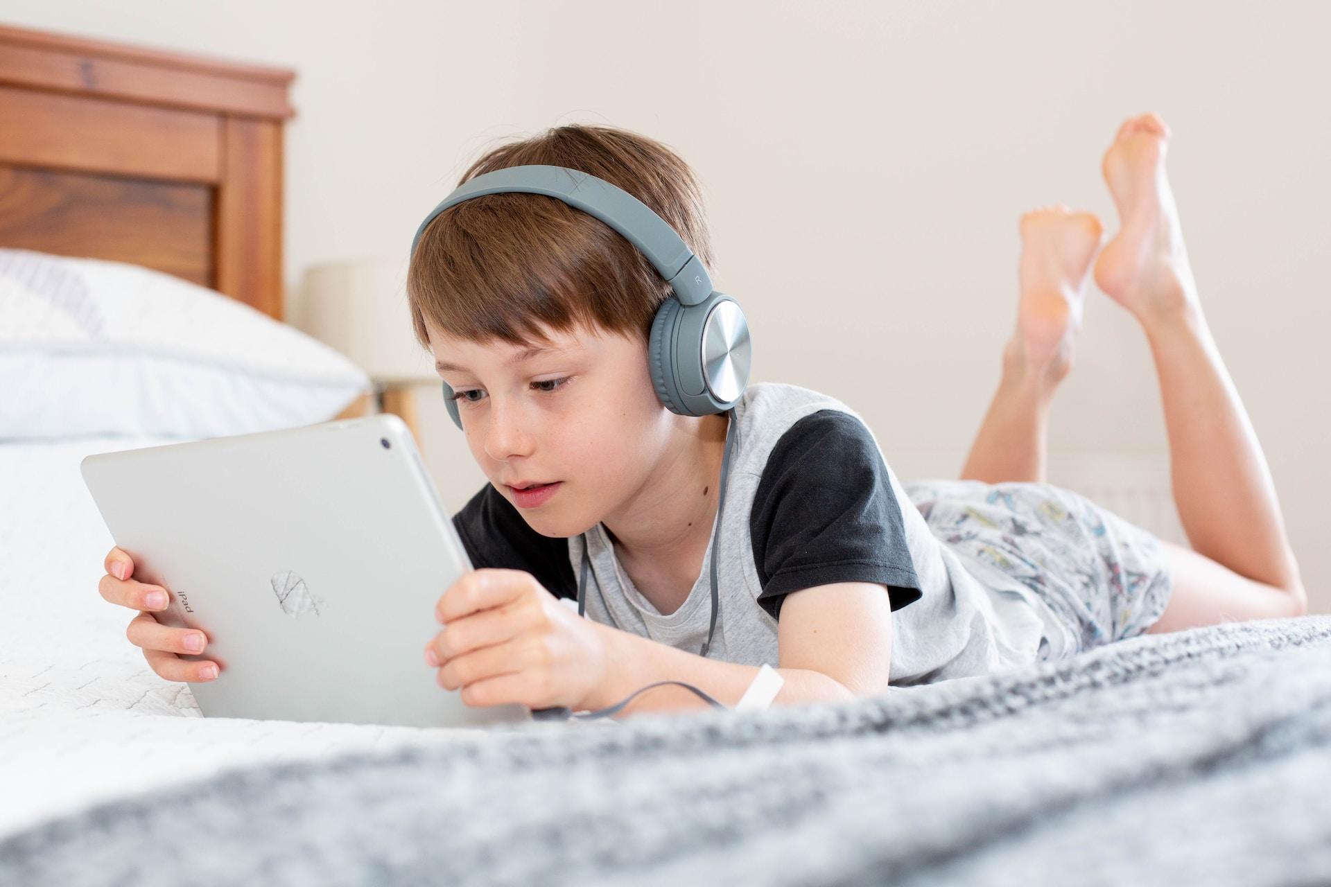 Gutt med t-skjorte og hodetelefoner ligger på senga og ser på en iPad.