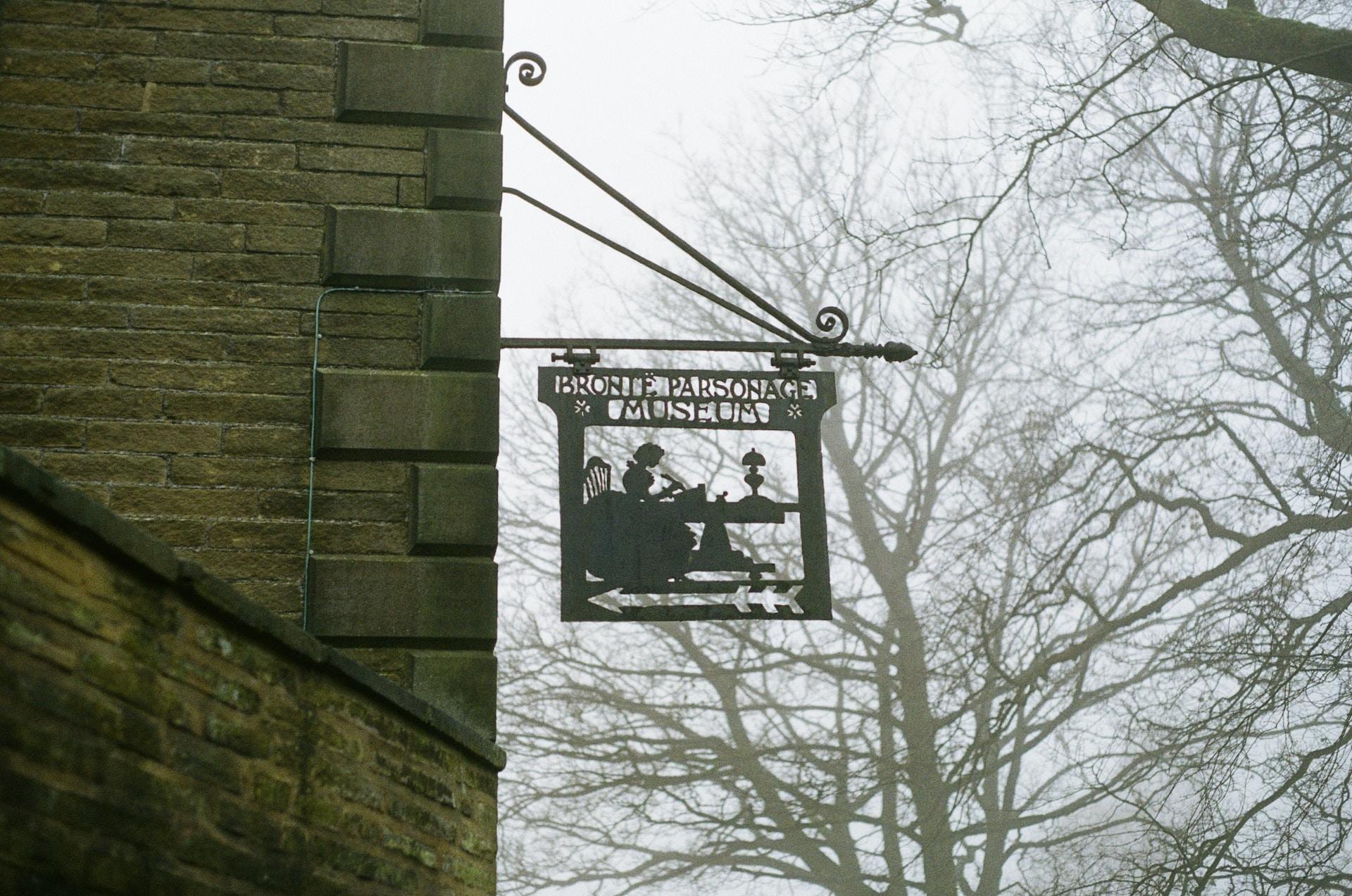 Metallskilt hengende ut fra murvegg med teksten "Brontë Parsonage Museum". 