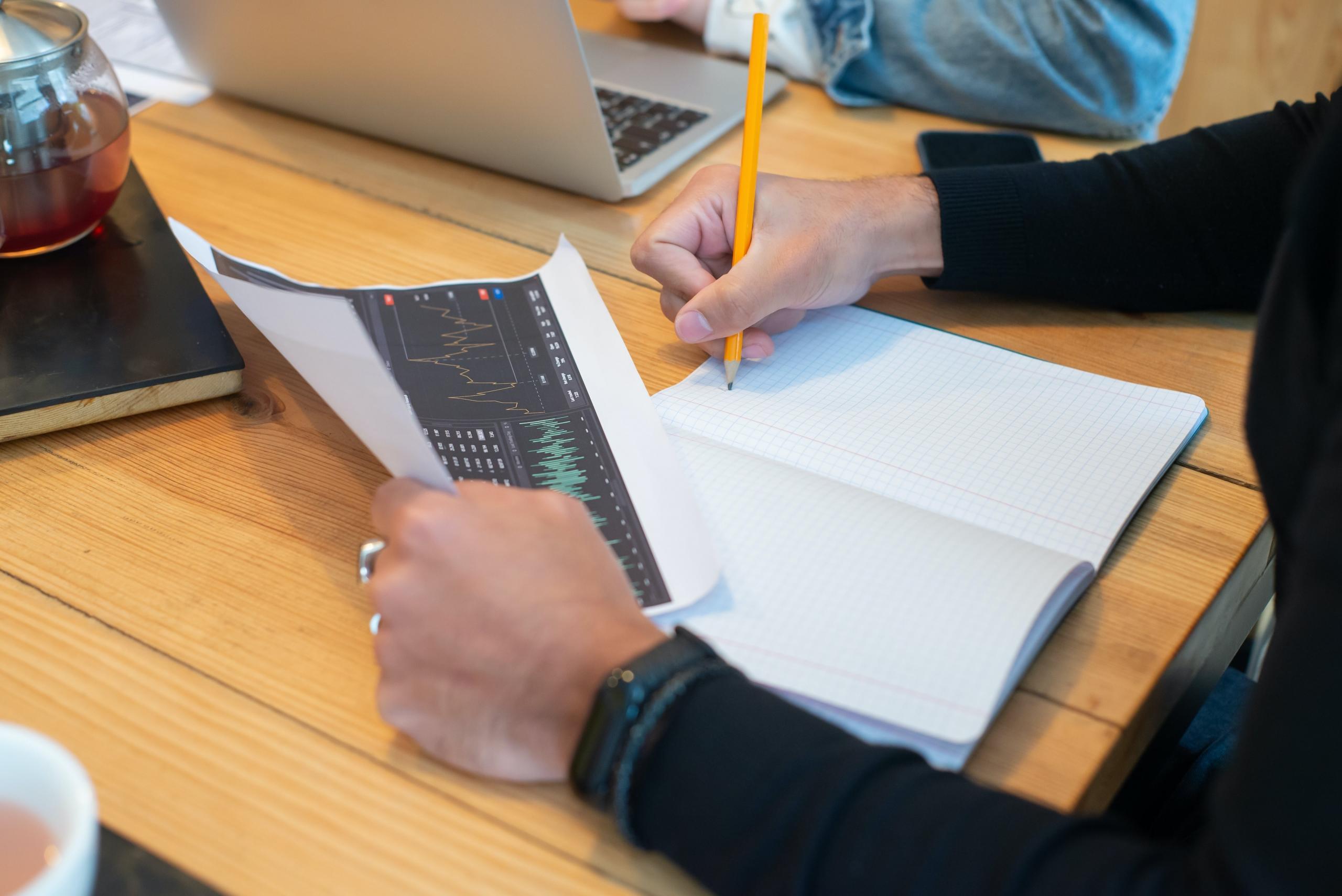 En person analyserer finansielle grafer og noterer nøkkelpunkter i en notatblokk ved siden av en bærbar datamaskin.