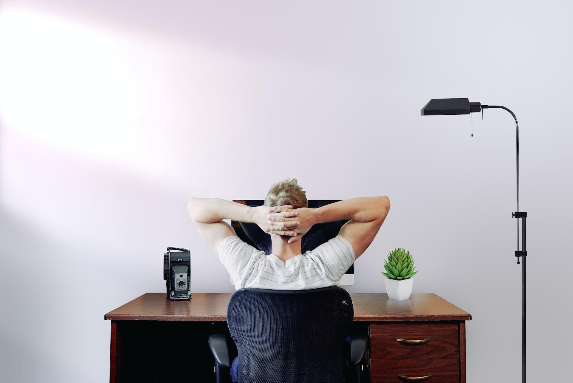Mann med blondt hår og hvit t-skjorte sitter på en svart kontorstol foran en PC-skjerm med hendene bak hodet.