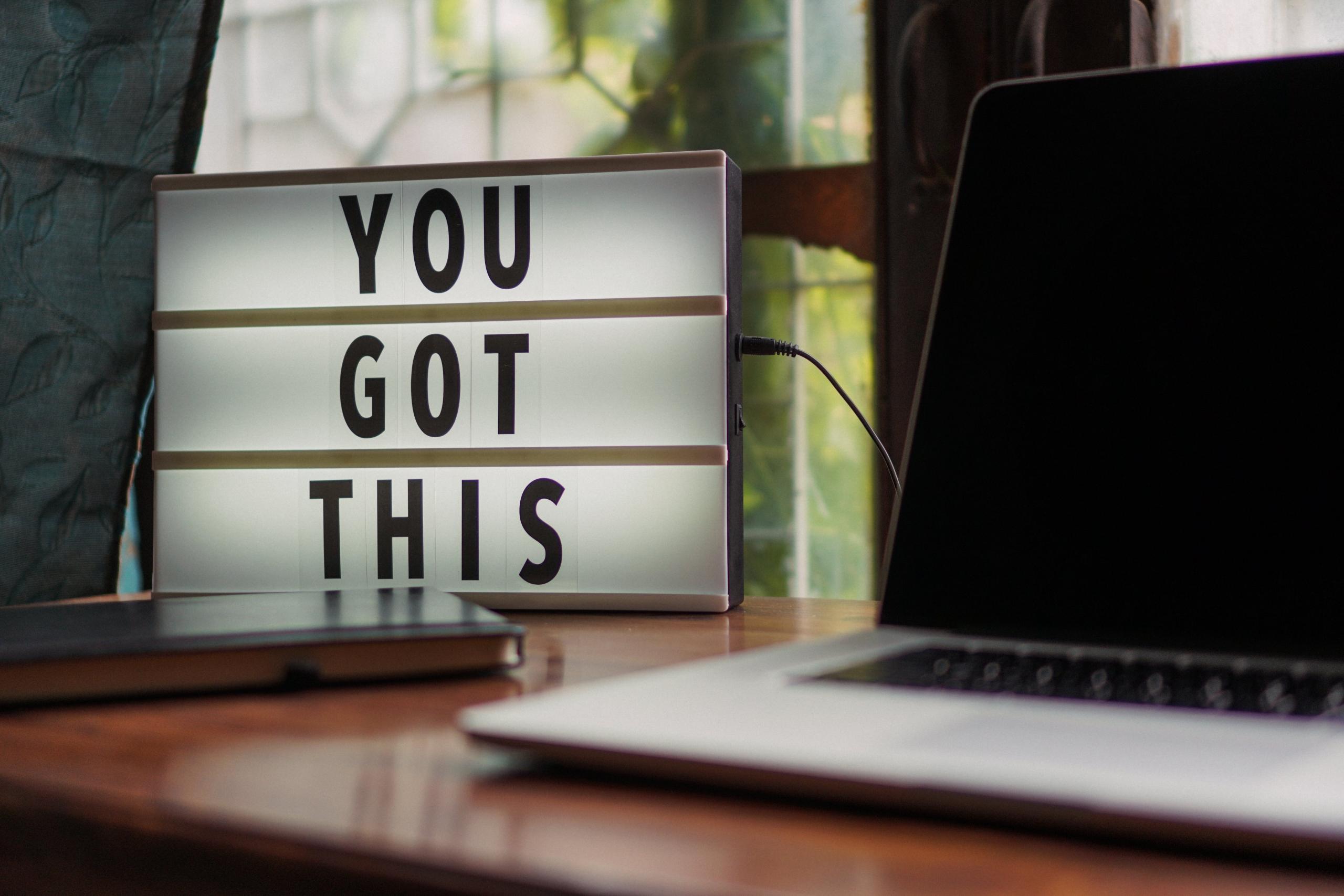 En lyskasse med teksten 'YOU GOT THIS' ved siden av en bærbar datamaskin på et skrivebord.