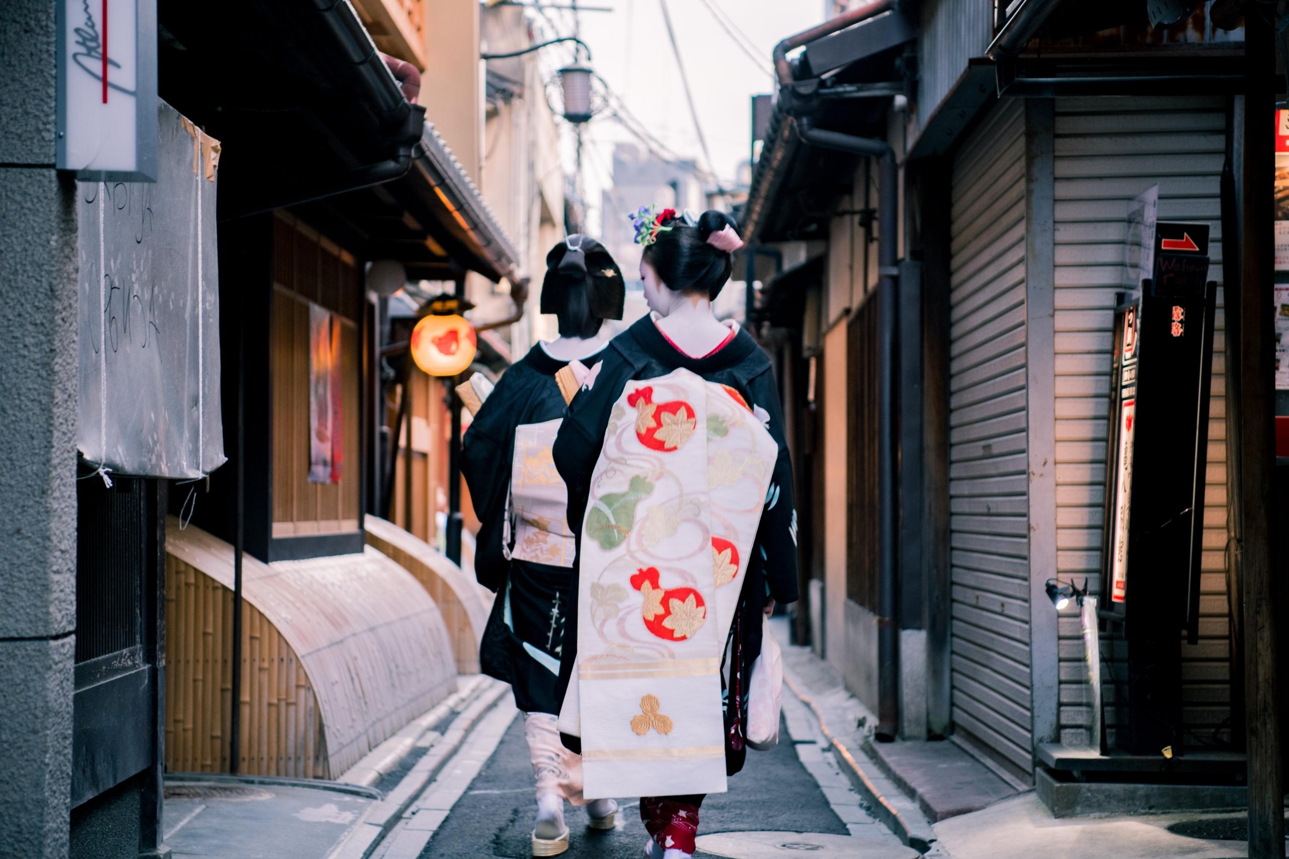 To geishaer går langs en smal vei i Kyoto, med ryggen vendt mot kameraet, viser de intrikate detaljene i deres kimonoer og hårornament.