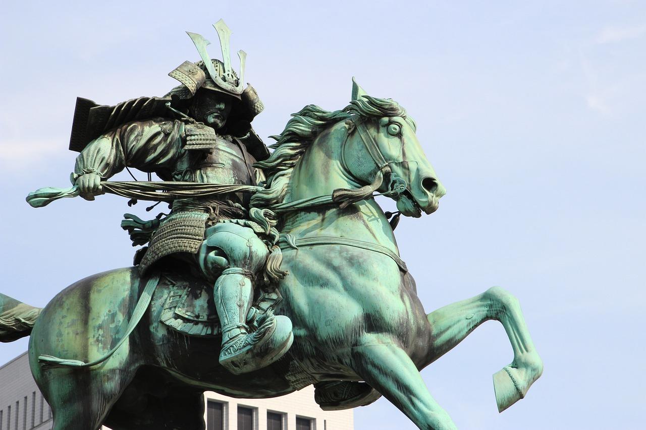 Bronsestatue av en samurai i full rustning på en hest som reiser seg på bakbena, mot en klar himmel.