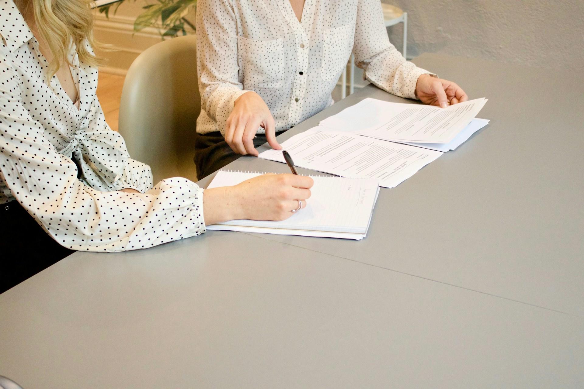 To kvinner med hvite bluser sitter ved siden av hverandre ved en pult og skriver på papirer.