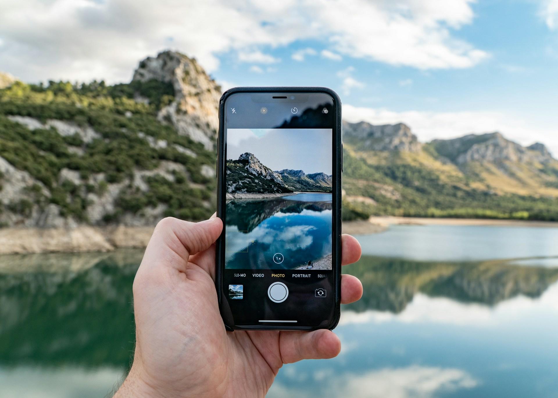 Hånd holder iPhone som tar bilde av en fjord med fjell.