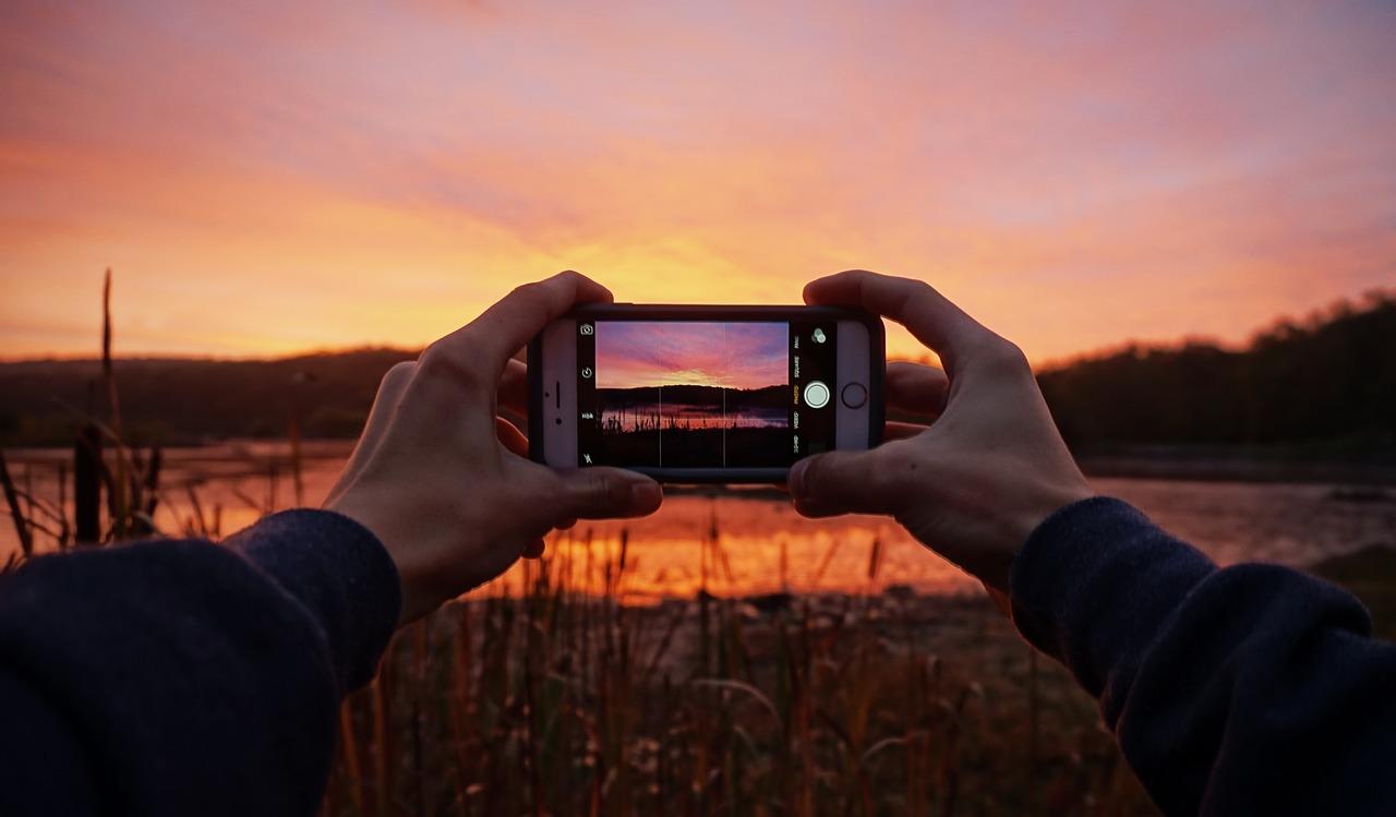 Hender som holder en smarttelefon som tar et bilde av en solnedgang over en elv.