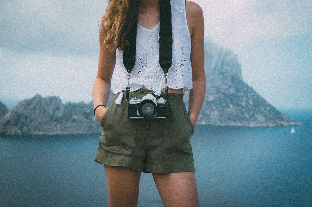 Kvinne med hvit bluse og olivengrønne shorts står med et vintage kamera rundt halsen foran et fjellandskap.