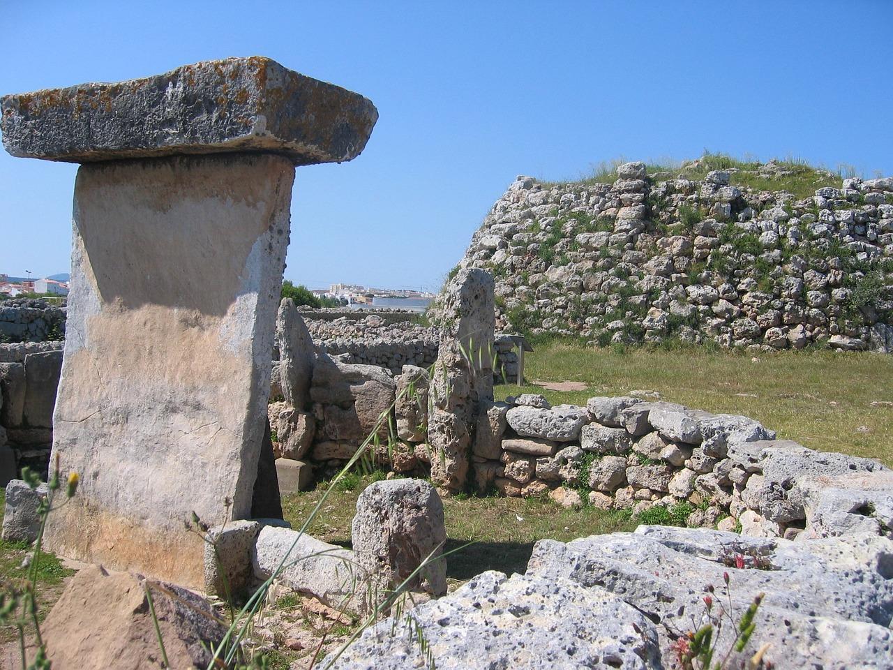 Det megalittiske monumentet Torralba d'en Salort på Menorca under en klar himmel, omgitt av ruiner og frodig vegetasjon.