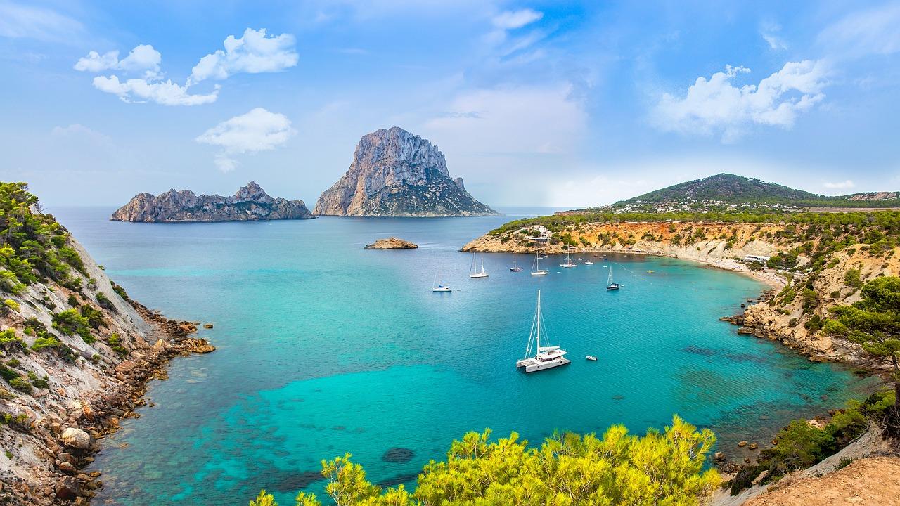Turkisblått vann og seilbåter i en idyllisk bukt ved Ibiza med den majestetiske Es Vedrà-klippen i bakgrunnen.