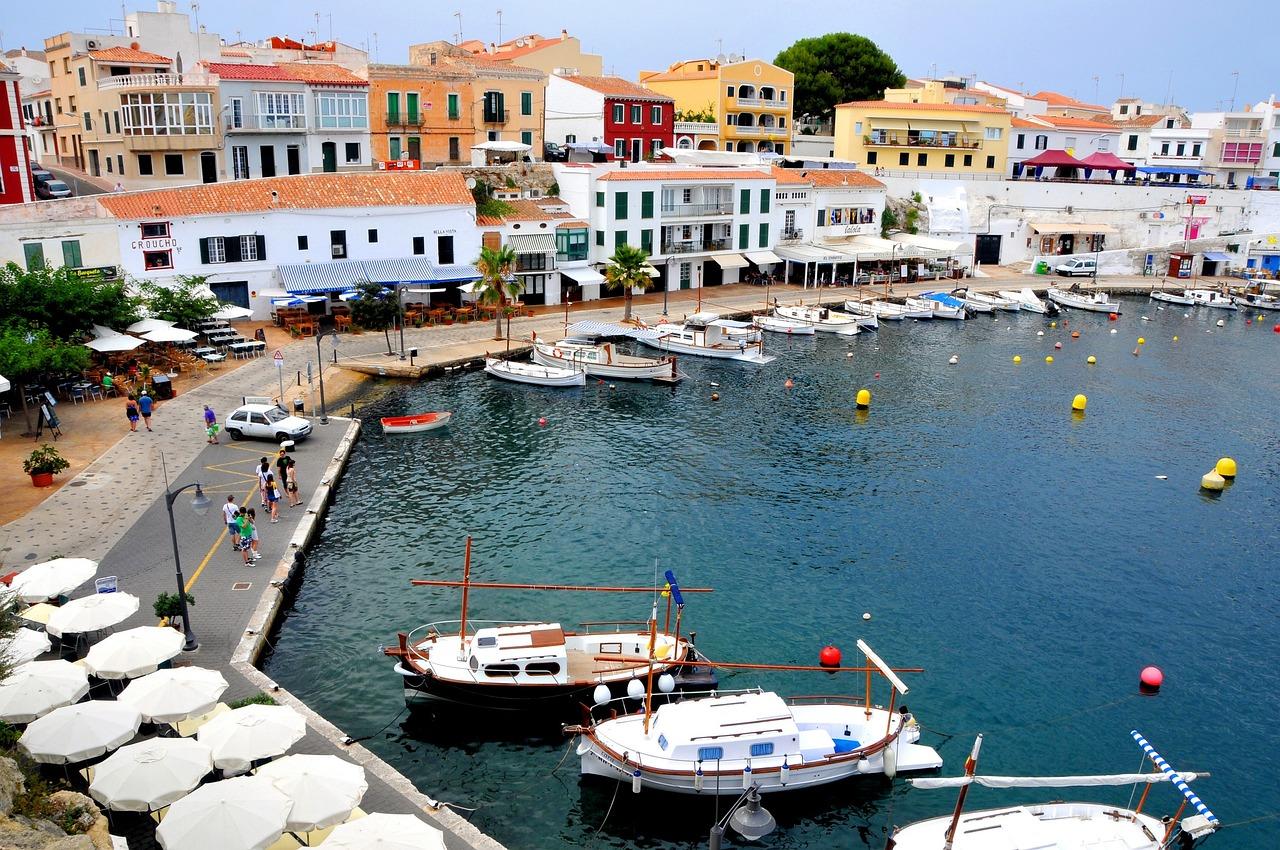 En fargerik havn i Menorca med tradisjonelle båter, omgitt av hvite bygninger med fargerike fasader og parasoller langs kaien.