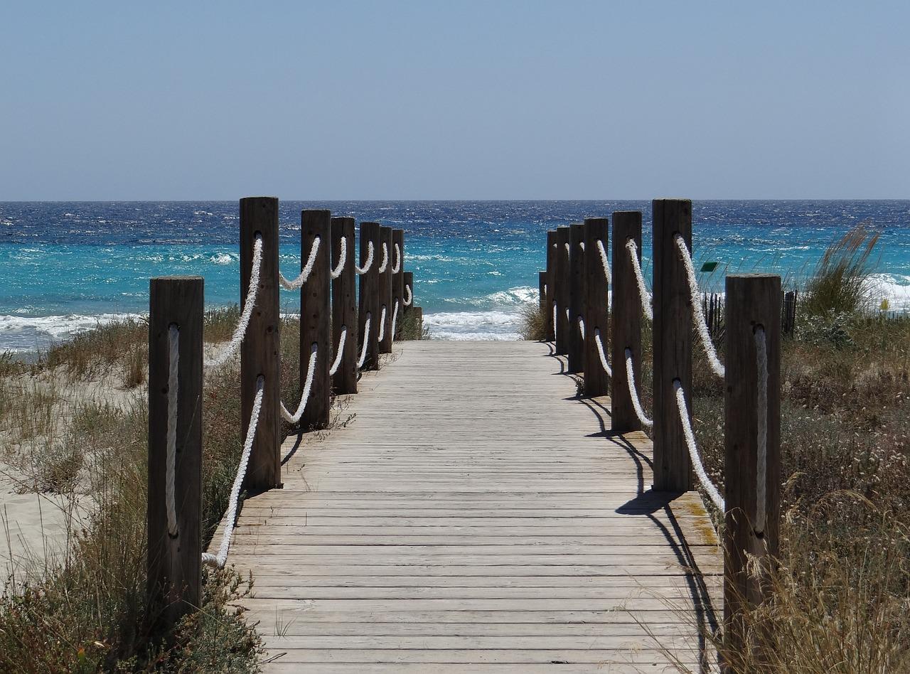 En trebro som leder til en innbydende strand på Menorca, med klart blått hav og sanddyner på sidene.