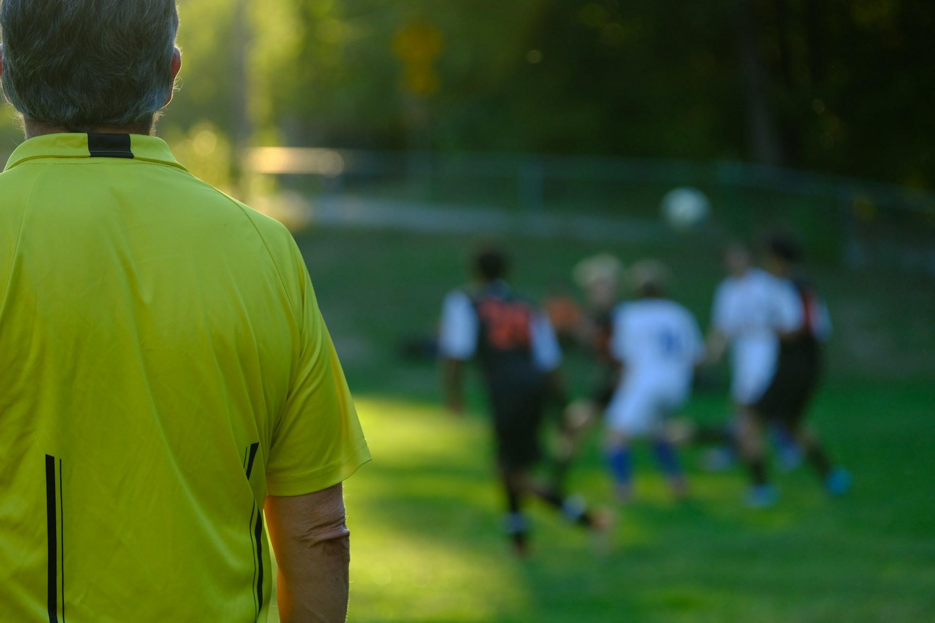 Mannlig fotballdommer med gul skjorte står på sidelinjen og følger nøye med på kampen.