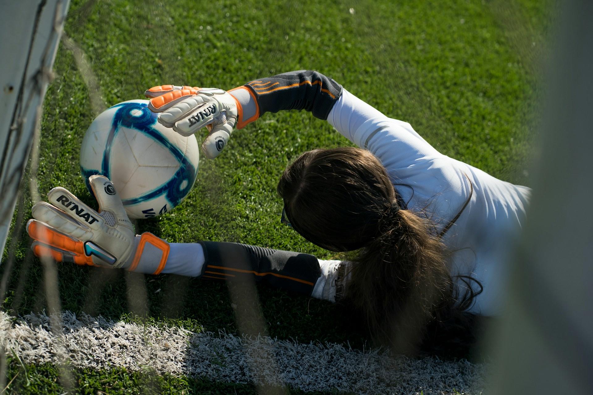 Kvinnelig keeper med langt brunt hår ligger på banen og har fanget fotballen med hendene.