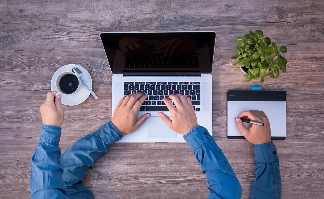 En person arbeider på en bærbar datamaskin mens han bruker en tegneplate, eksempel på multitasking på arbeidsplassen.