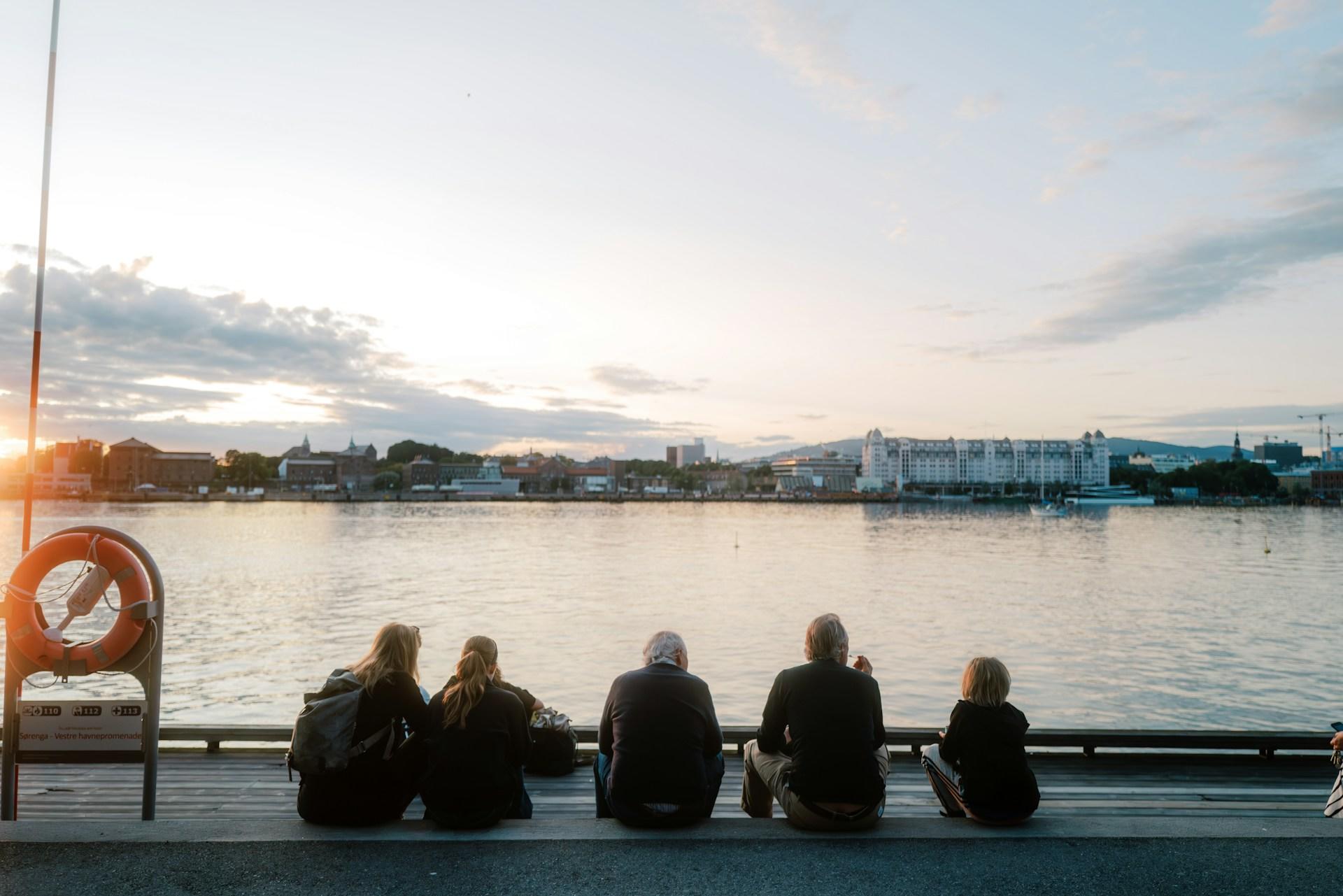 Mennesker sitter og ser utover vannet i Oslo, på brygga.