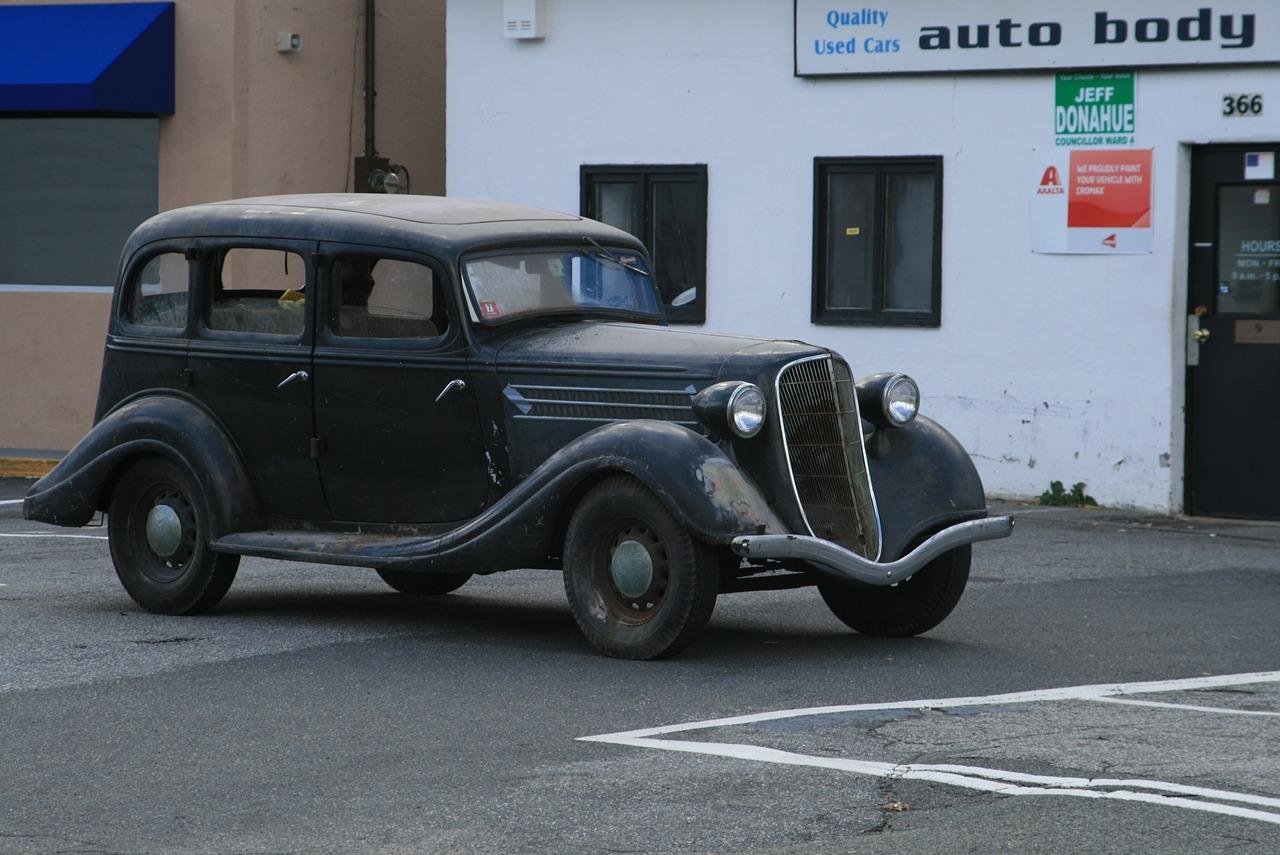 En svart bil fra 1930-tallet står parkert foran et bilverksted.