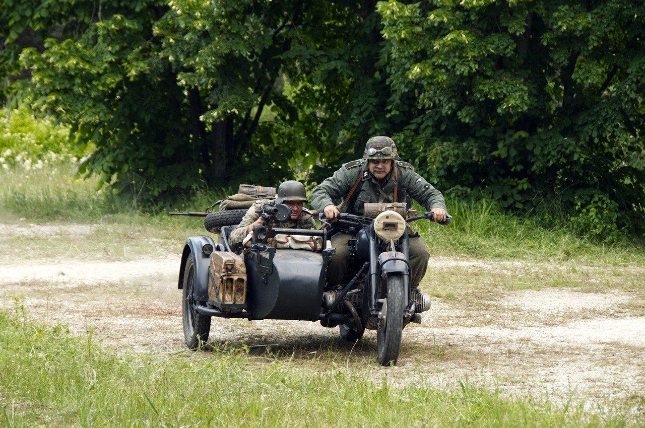 To soldater i tyske uniformer under andre verdenskrig kjører en motorsykkel med sidevogn gjennom en skogkledd sti.