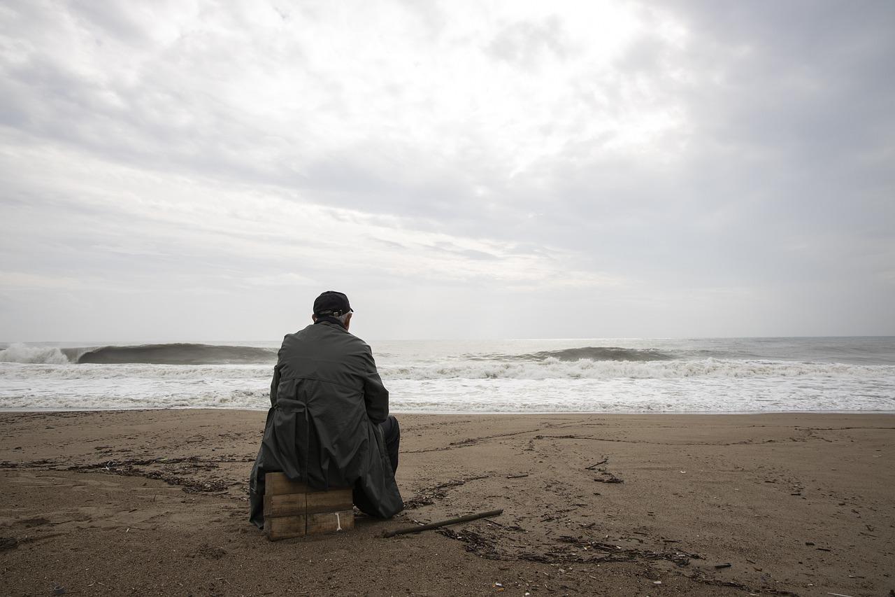 En mann sitter alene på en trekasse ved stranden og ser utover havet, symboliserer isolasjon og håpløshet.