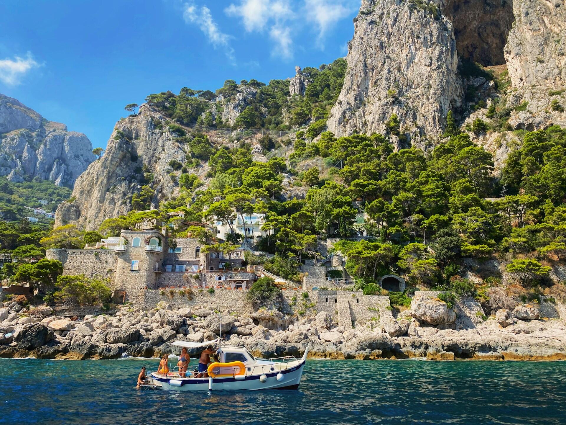 Liten båt med turister ved klipper og villaer langs kysten av Capri Itali