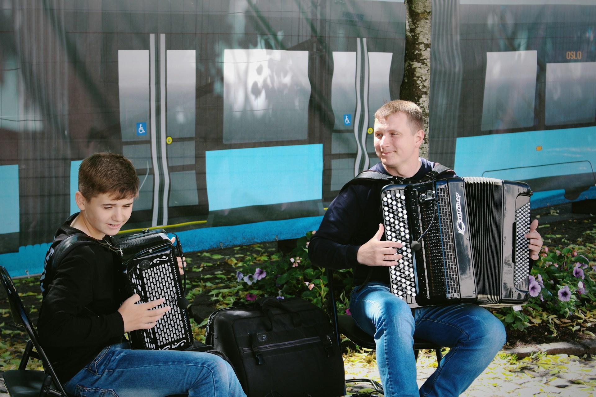 Far og sønn spiller på hvert sitt trekkspill med blomster bak.