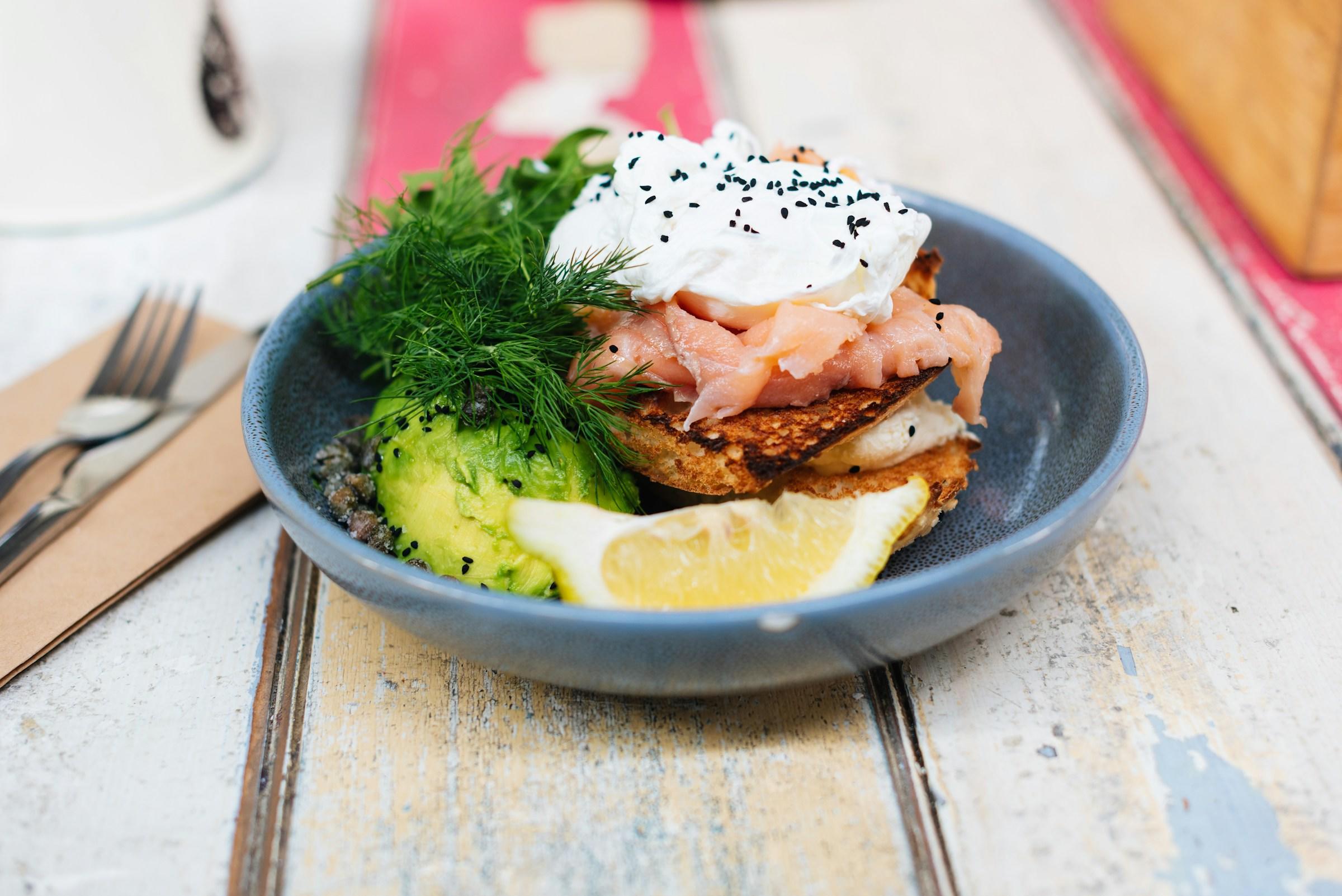 En blå tallerken på et trebord med et smørbrød på. Smørbrød med laks, egg og avocado.