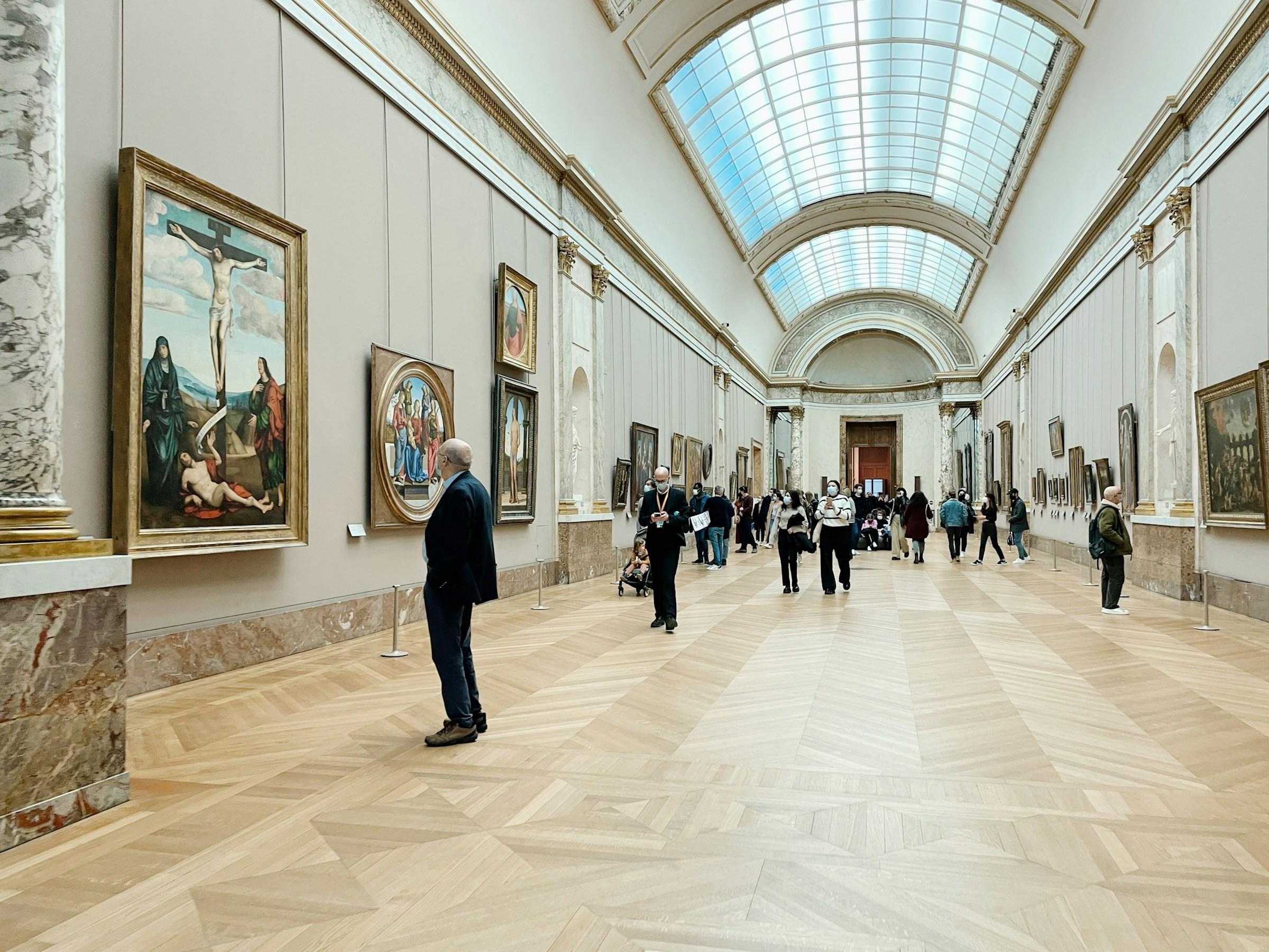 Bilde tatt fra Louvre museumet. Flere mennesker står i et rom og observerer kunst på veggene.