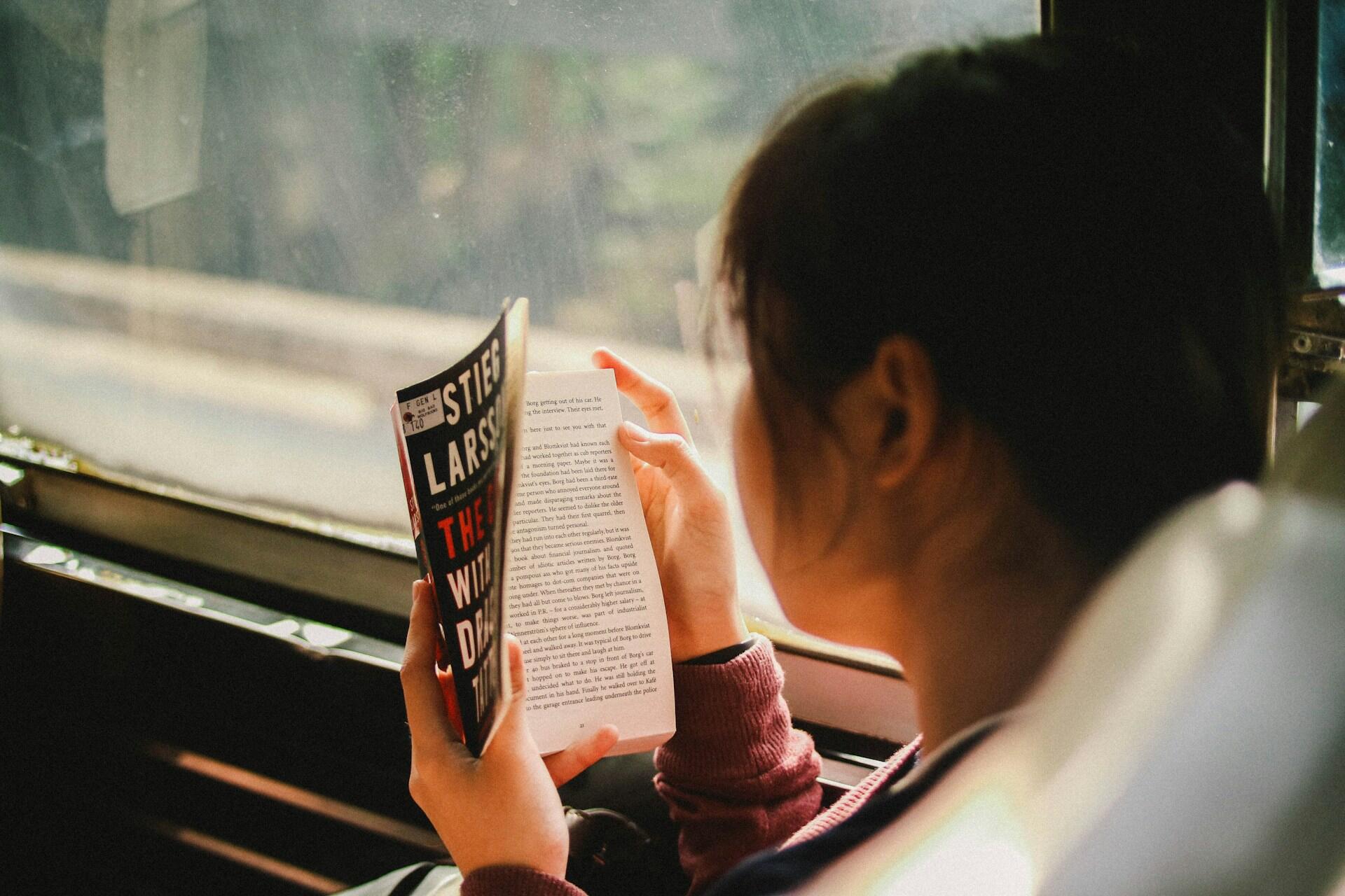 Kvinne leser en bok mens hun sitter på en buss, med utsikt gjennom vinduet.