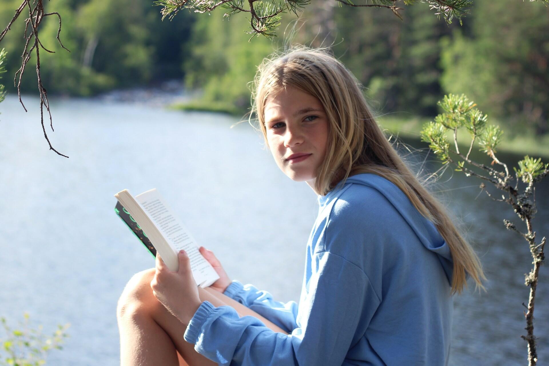 Ung jente leser en bok ute i naturen, ved en innsjø.