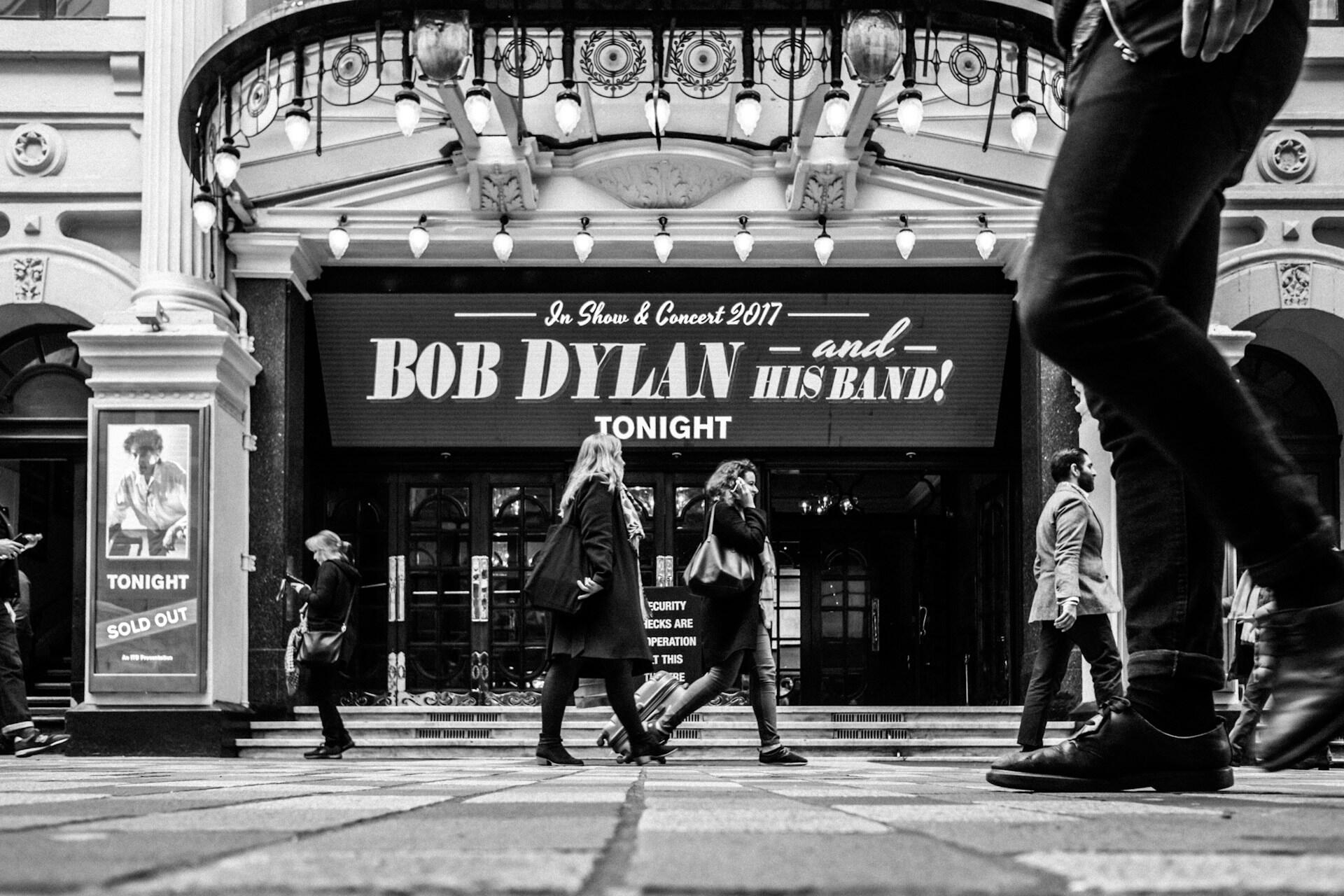 Folk går forbi teater med plakat for Bob Dylan-konsert, «Sold Out» og 2017-markering.