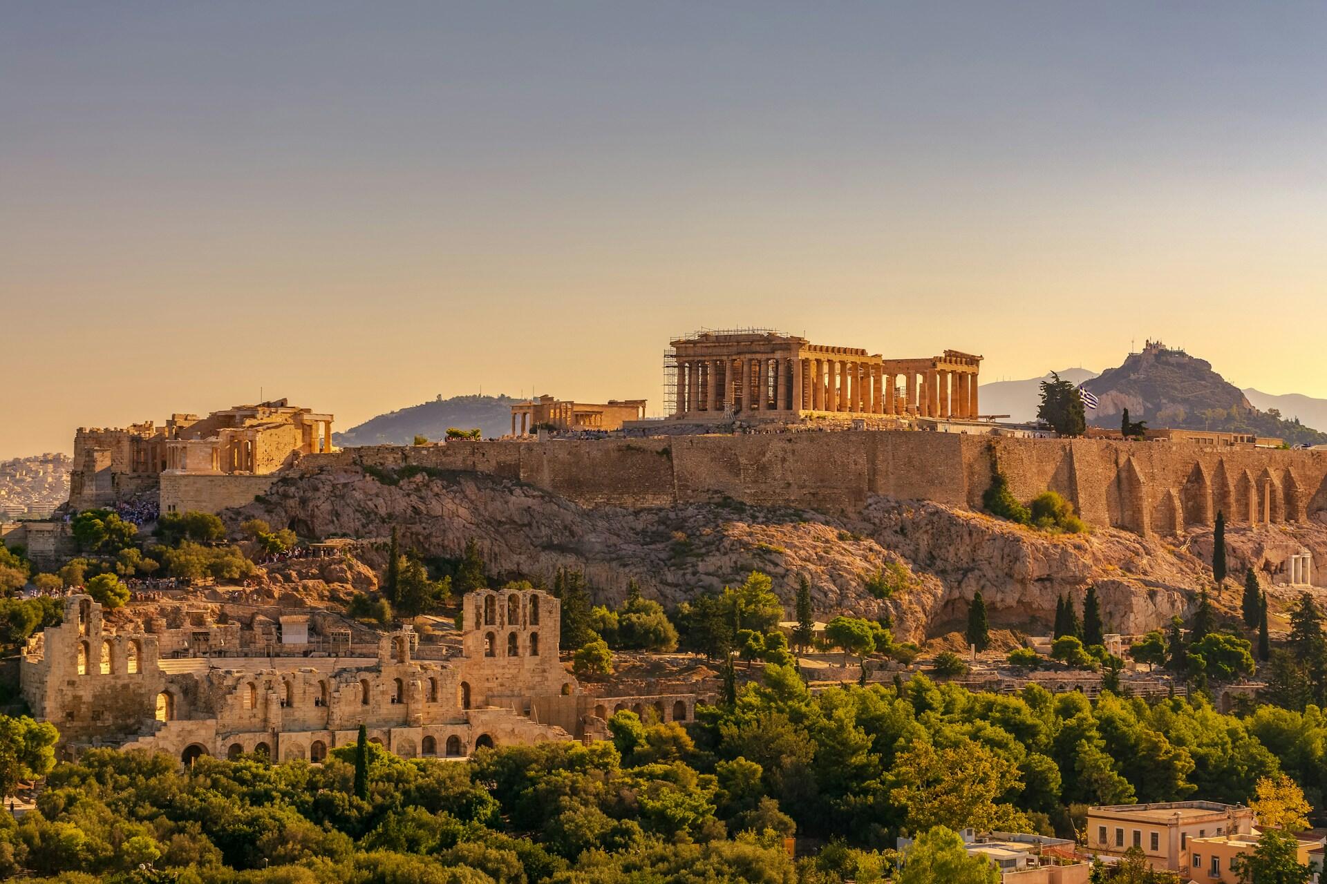 Akropolis-bygningen på toppen av en høyde i Athen, med gamle bebyggelser rundt og solnedgang i bakgrunnen.