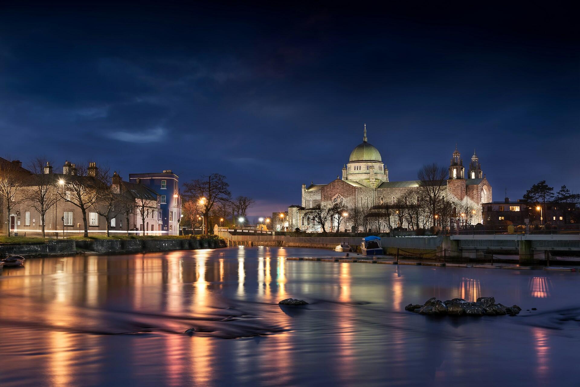 Galway Cathedral ved elven Corrib i skumringen, med lys som speiler seg i vannet