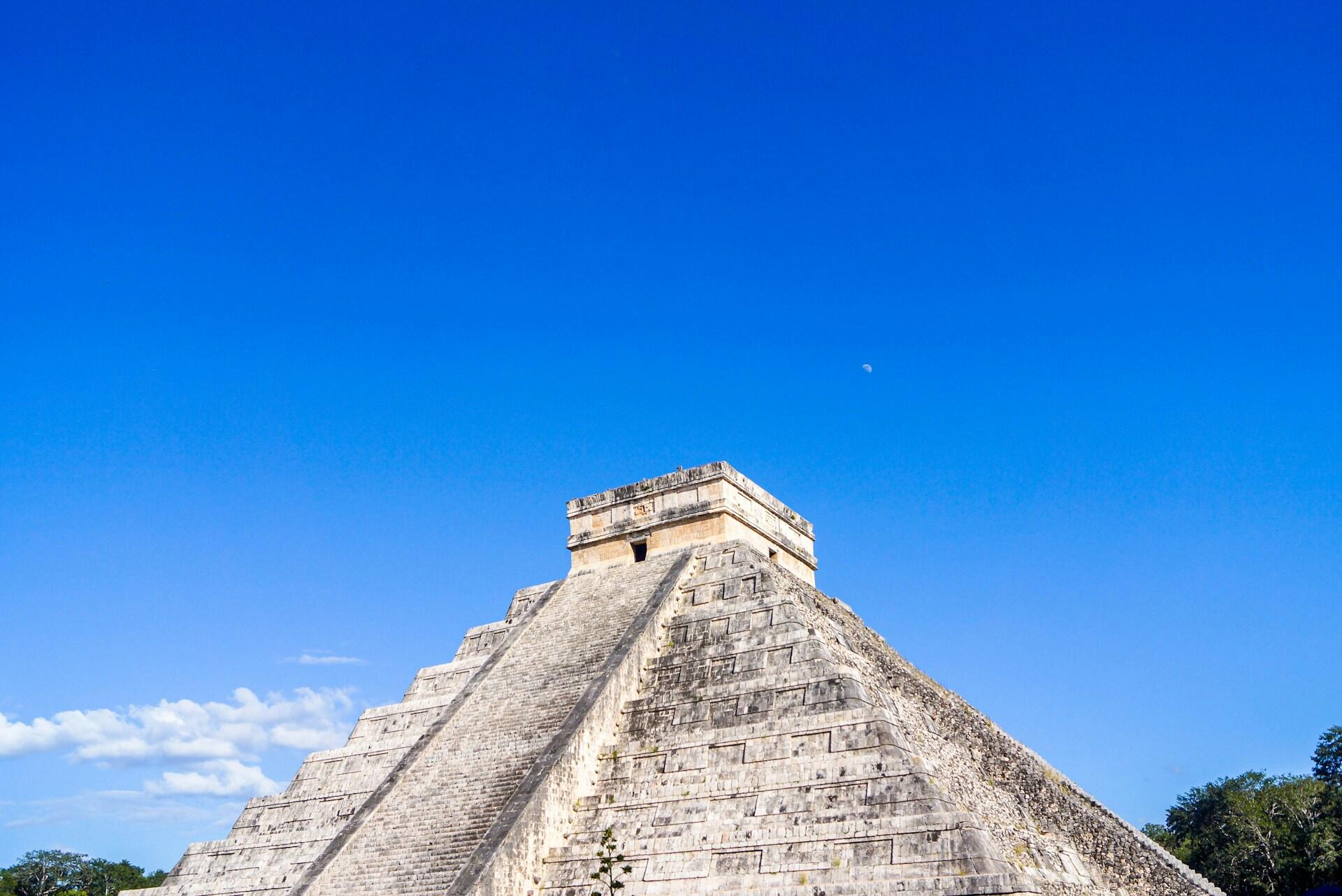 Maya-tempel laget av grå stein med grønne, frodige trær og blå himmel i bakgrunnen.
