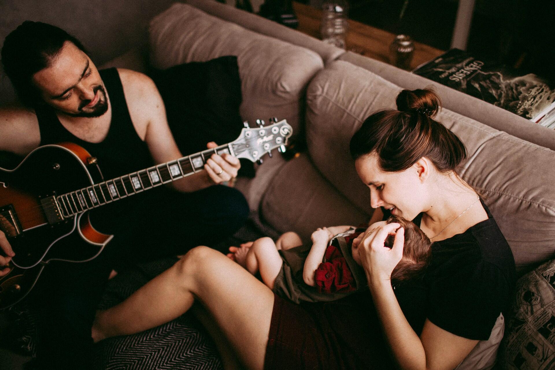 Far spiller gitar mens mor holder babyen i sofaen, i en intim og varm familiestund.