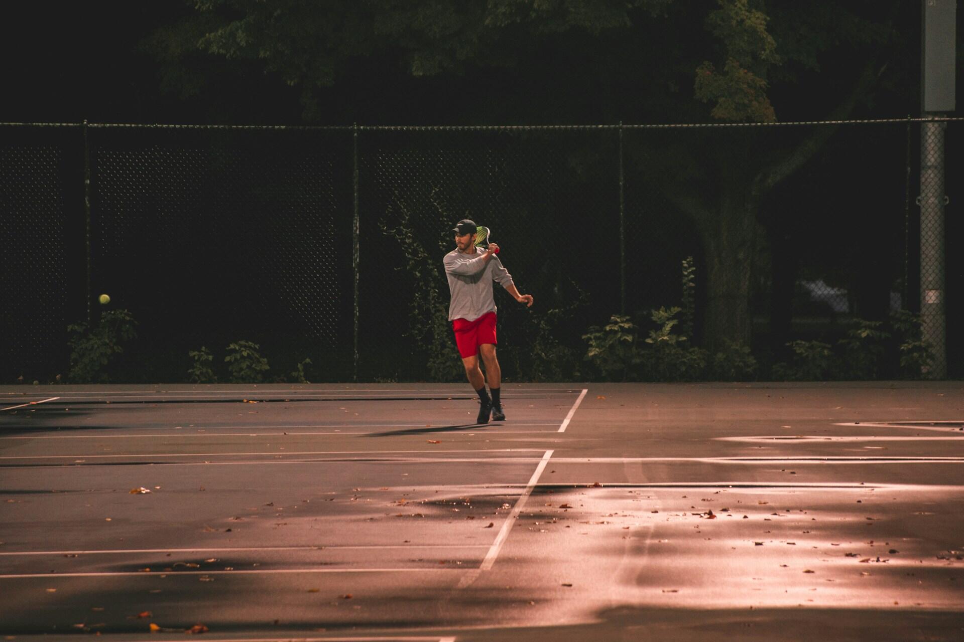 Tennisspiller i ferd med å slå ballen på en utendørs bane i skumringen.