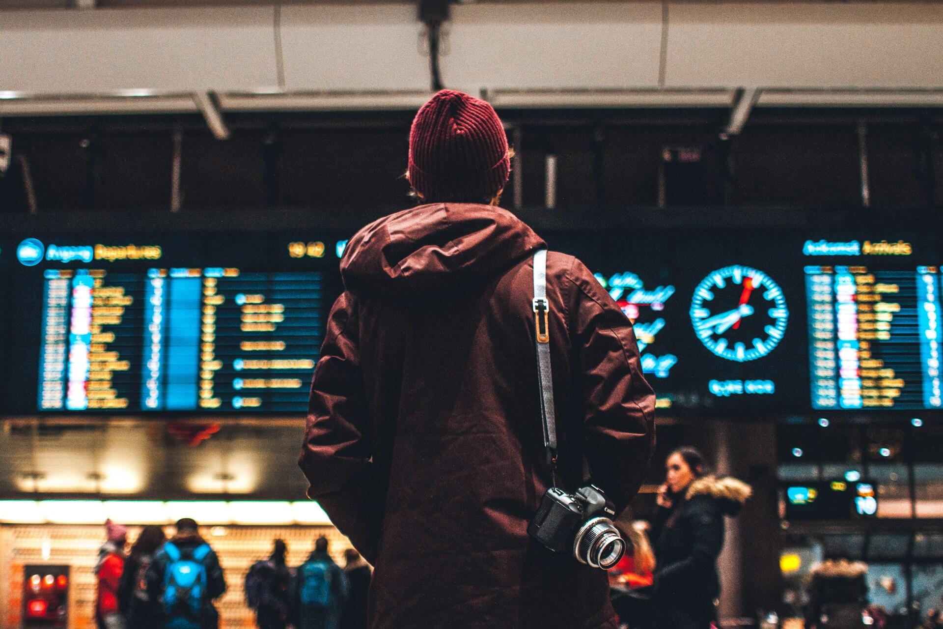 Mann med rød lue og jakke og kamera står med ryggen til og ser på tavle med info på flyplass.