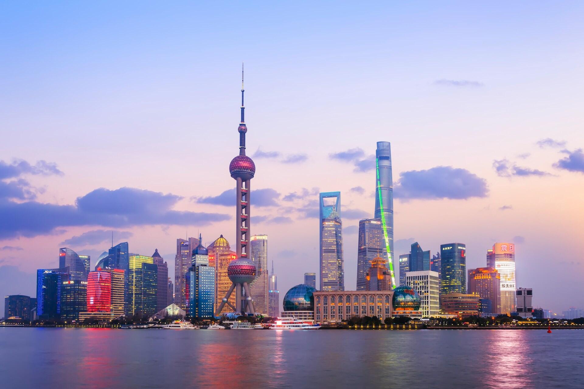 Den kinesiske byen Shanghai opplyst i mange ulike farger på kveldstid, med elven i forgrunnen.