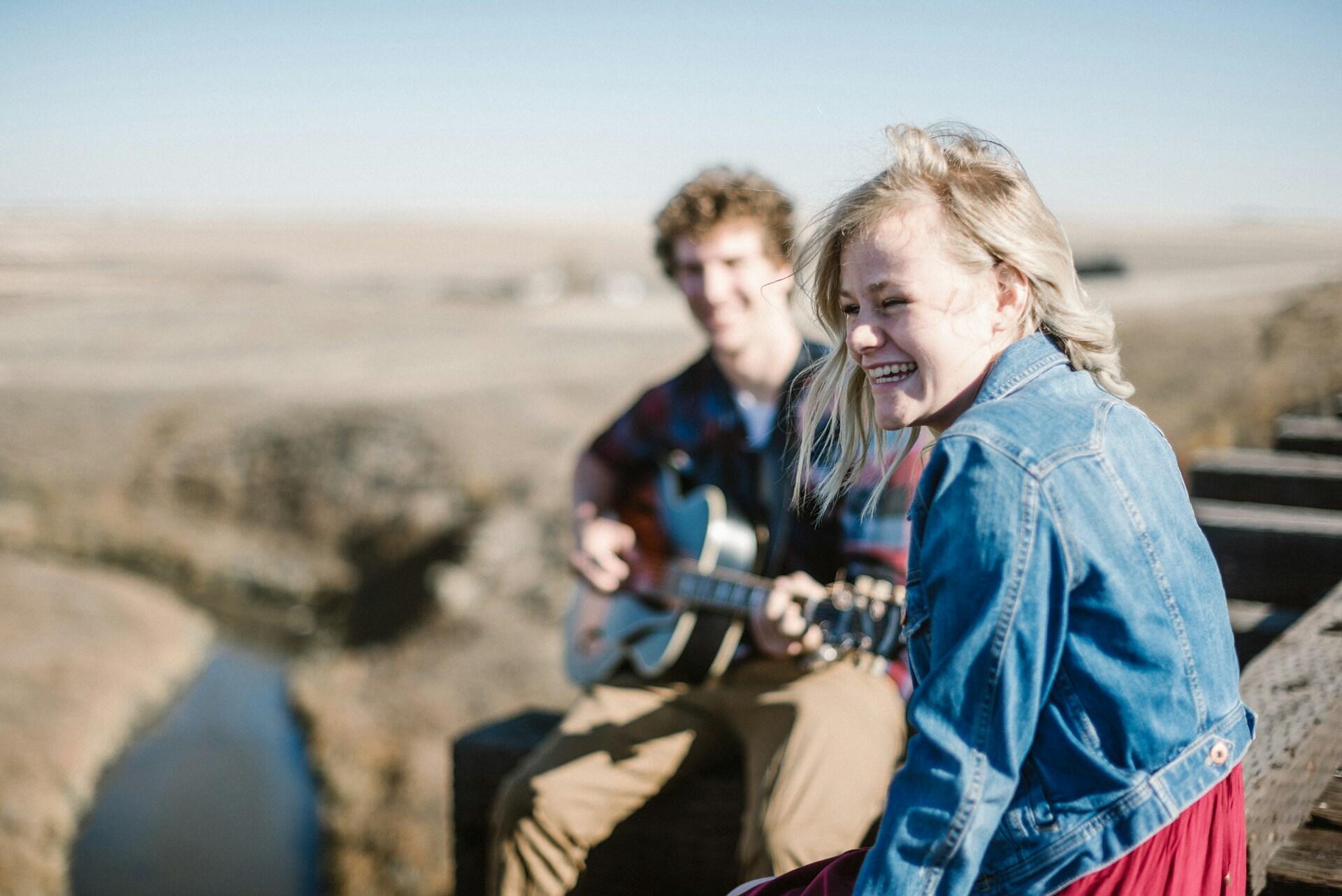 To unge personer smiler mens en av dem spiller gitar ute i naturen.